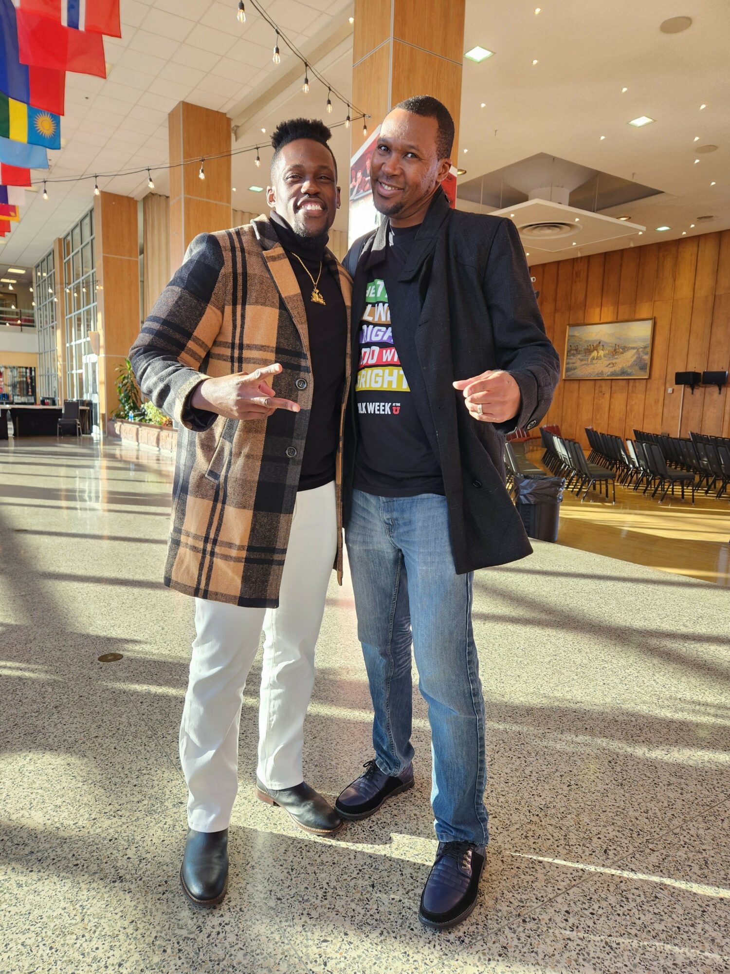 Two people standing in an indoor space with wooden walls and a tiled floor, one holding a phone, both dressed casually.