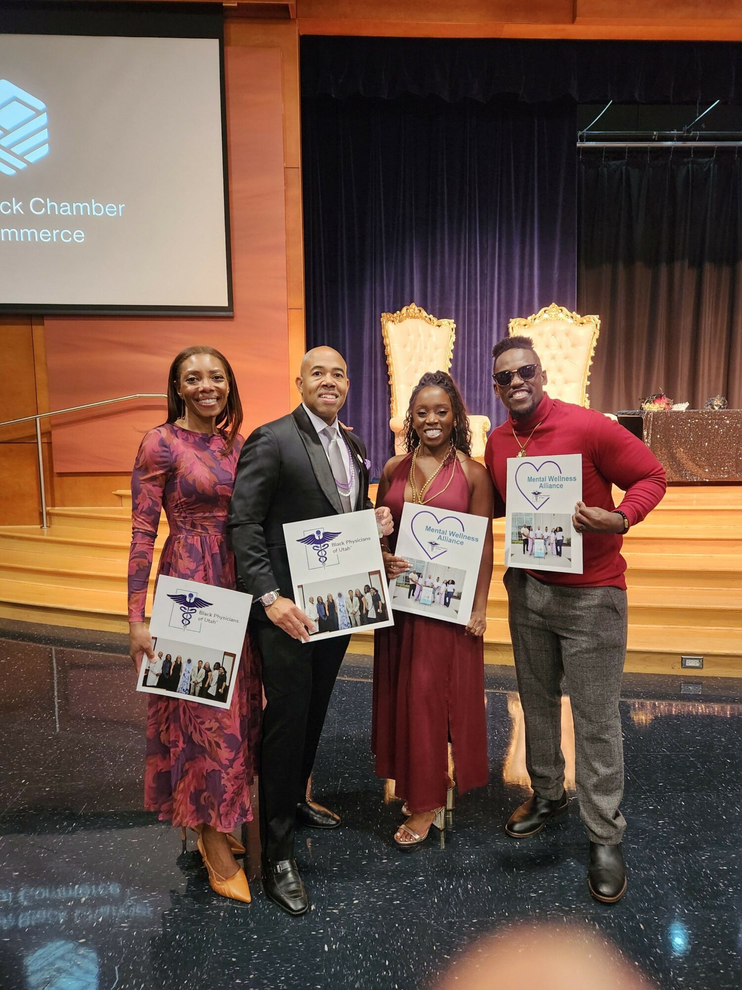 Three people standing on stage holding certificates, with a curtain and a screen in the background.