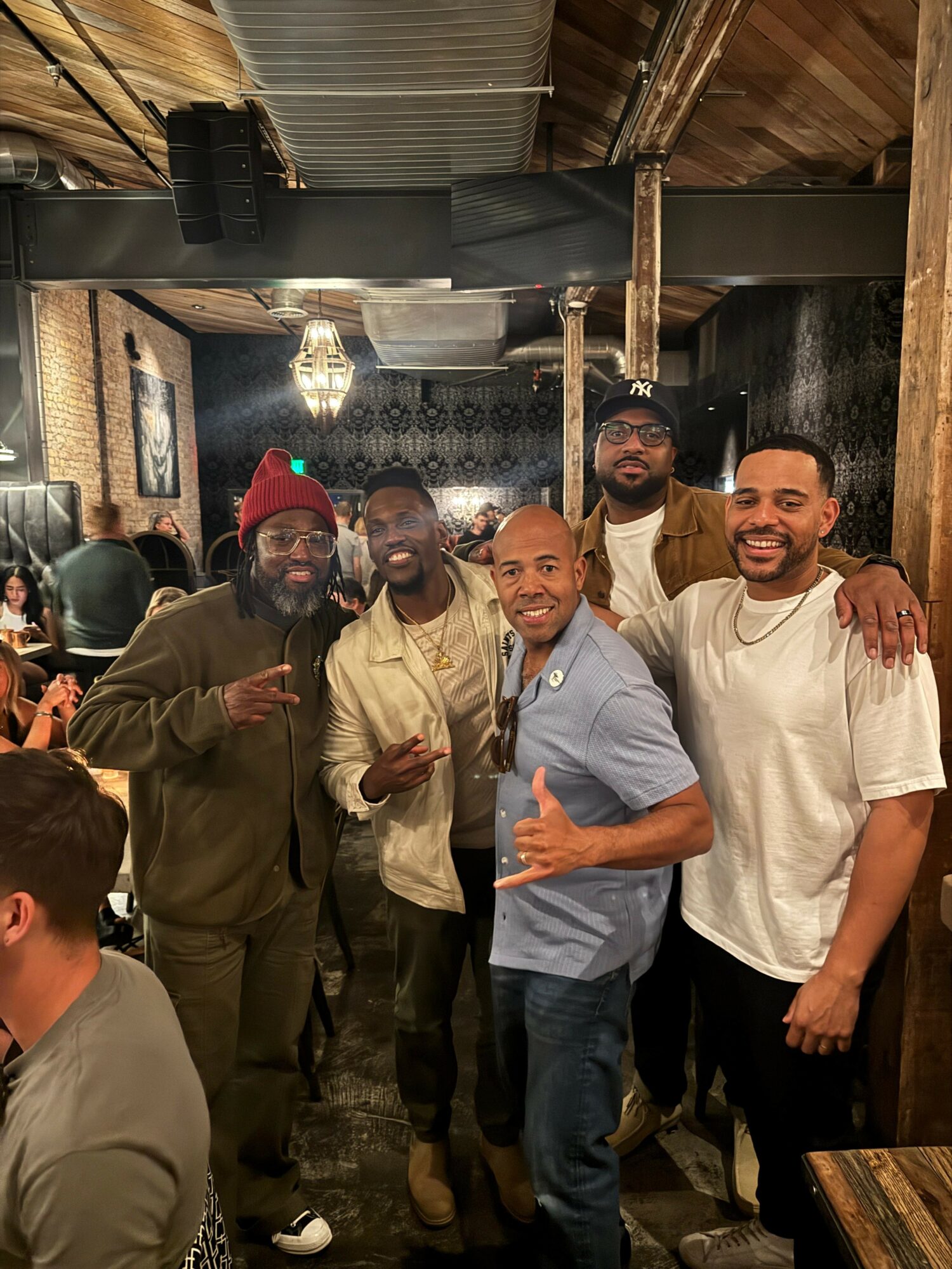 Five men standing together in a restaurant, smiling and posing for the photo, with a wooden ceiling and decorative lighting.