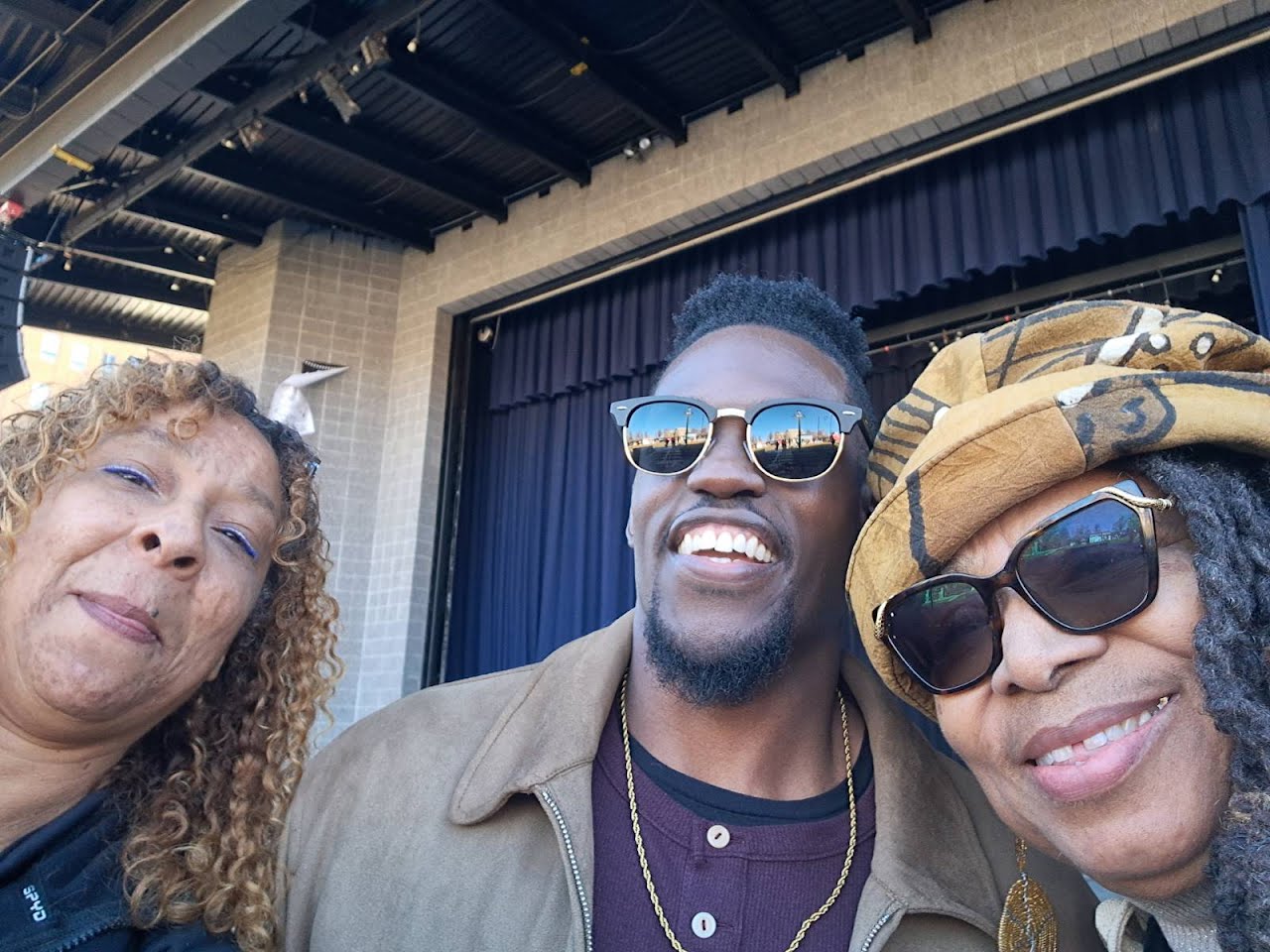 Three people smiling outdoors, wearing sunglasses, with a stage and curtain in the background.