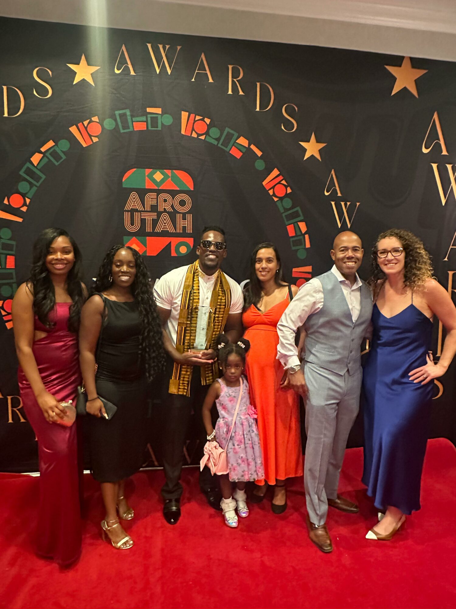 Group of seven people standing on red carpet in front of a black backdrop with gold stars and text, smiling for photo.