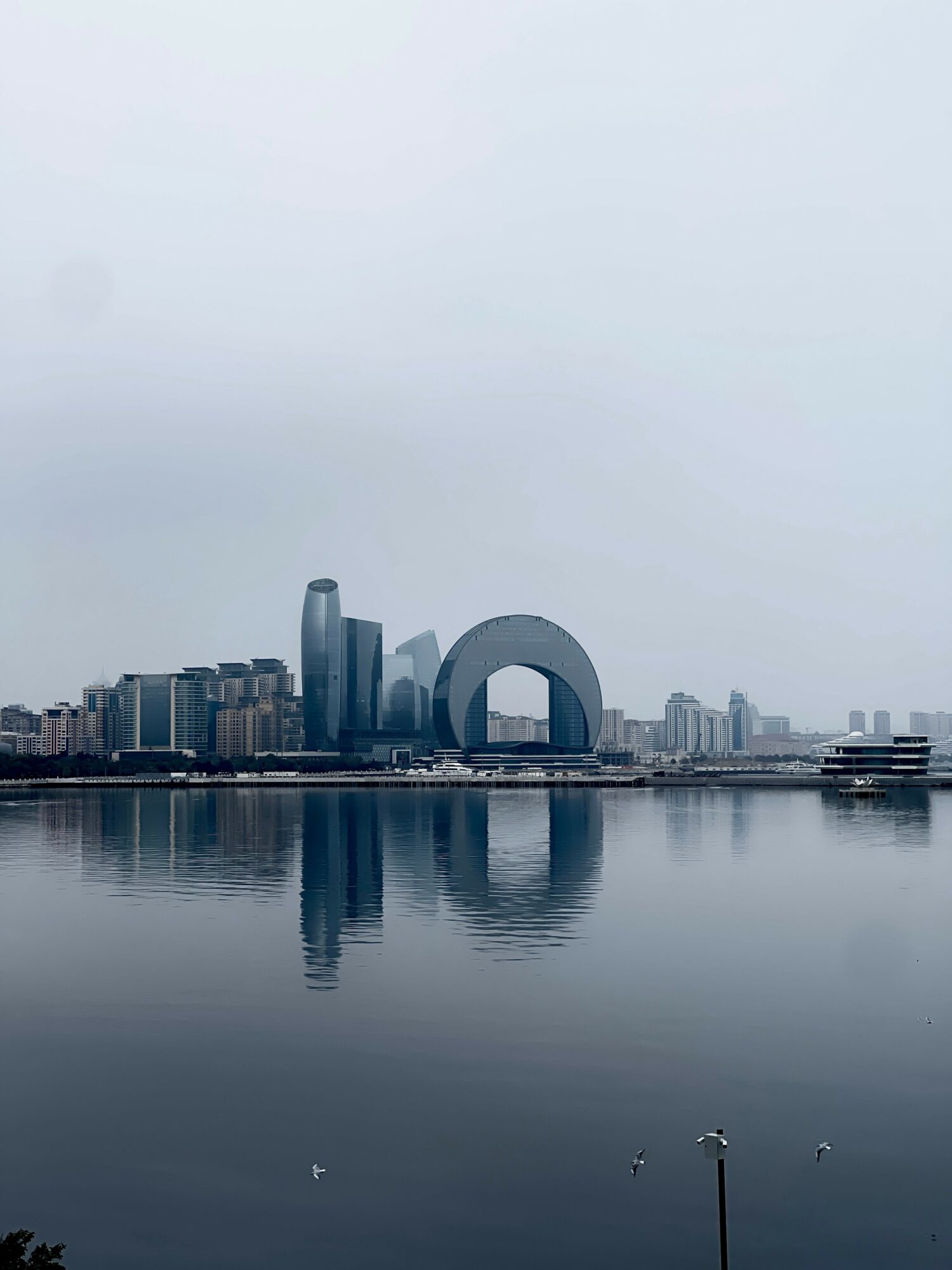 City skyline with modern buildings reflected in water, under cloudy sky, with a large circular structure in the center.