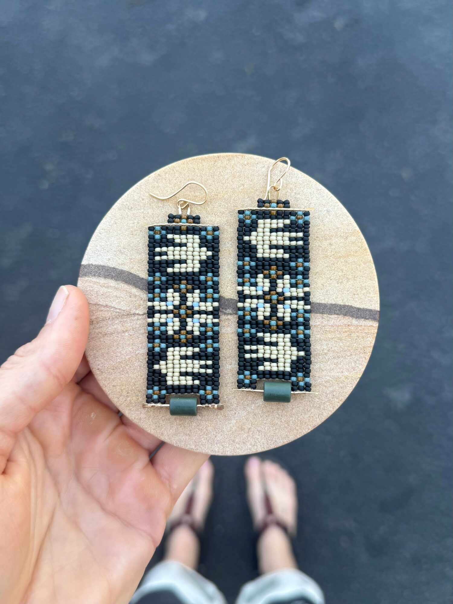 Hand holding circular display with beaded rectangular earrings against dark background.