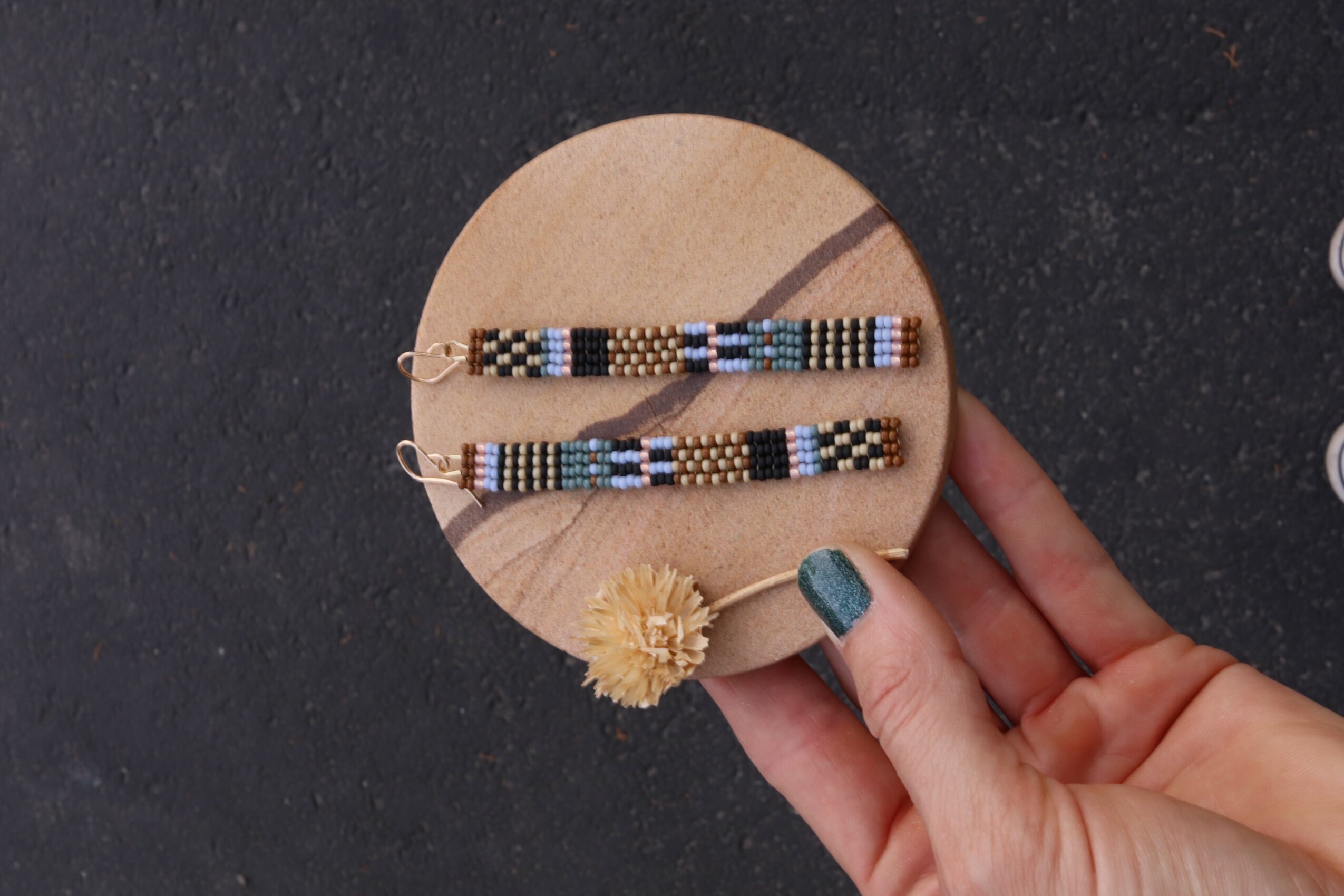 Hand holding a round wooden board with beaded jewelry and a flower-shaped accessory, dark background.