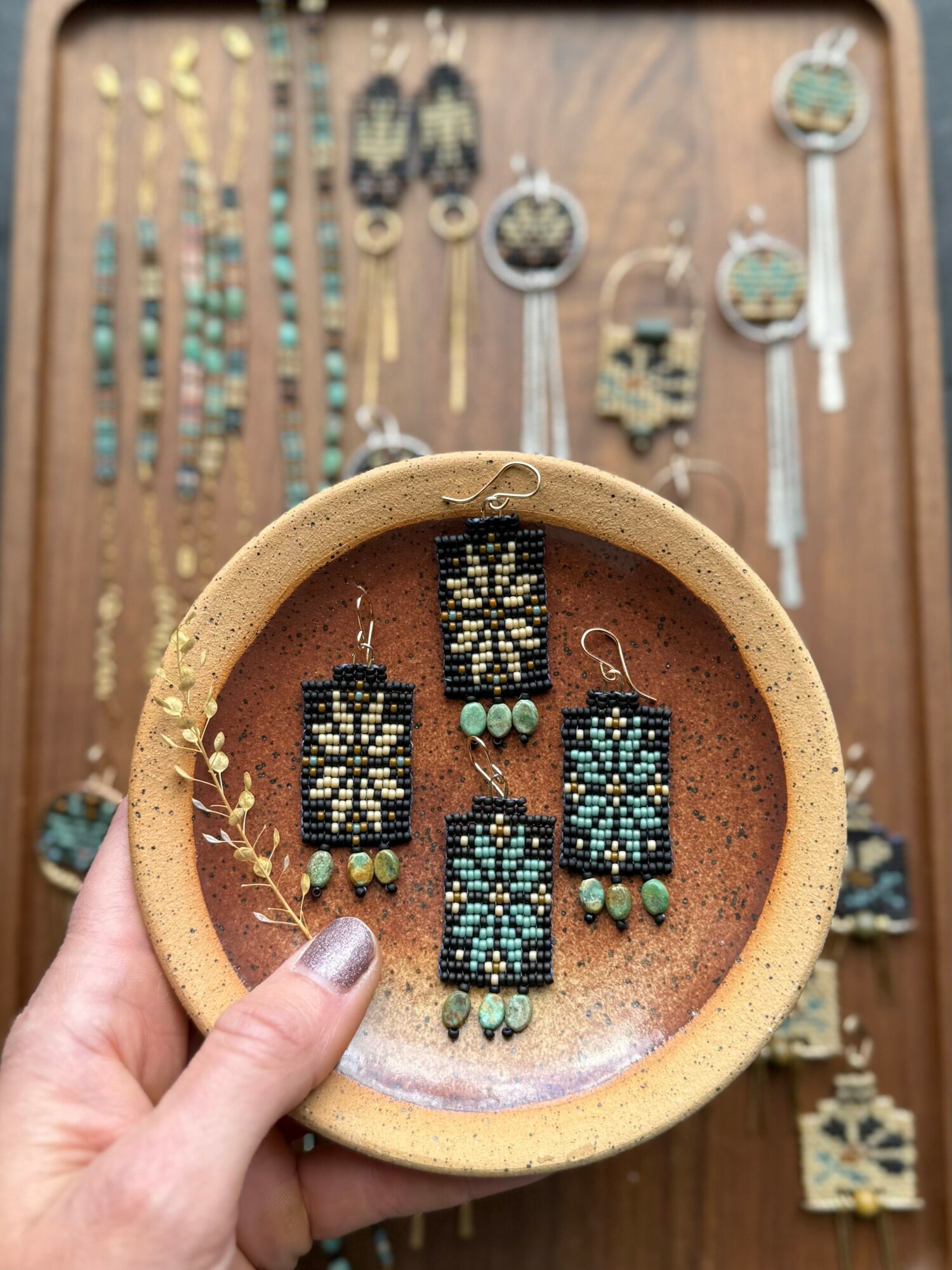 Hand holding a round dish with beaded jewelry, with more jewelry displayed on a wooden background behind.