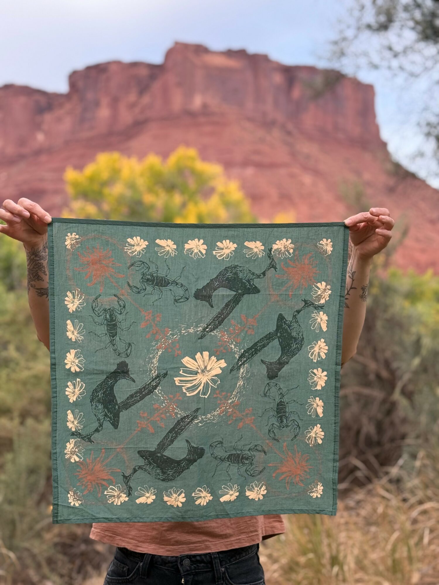 Person holding a decorated cloth with guitars, flowers, and leaves, outdoors with a red rock formation and trees in background.