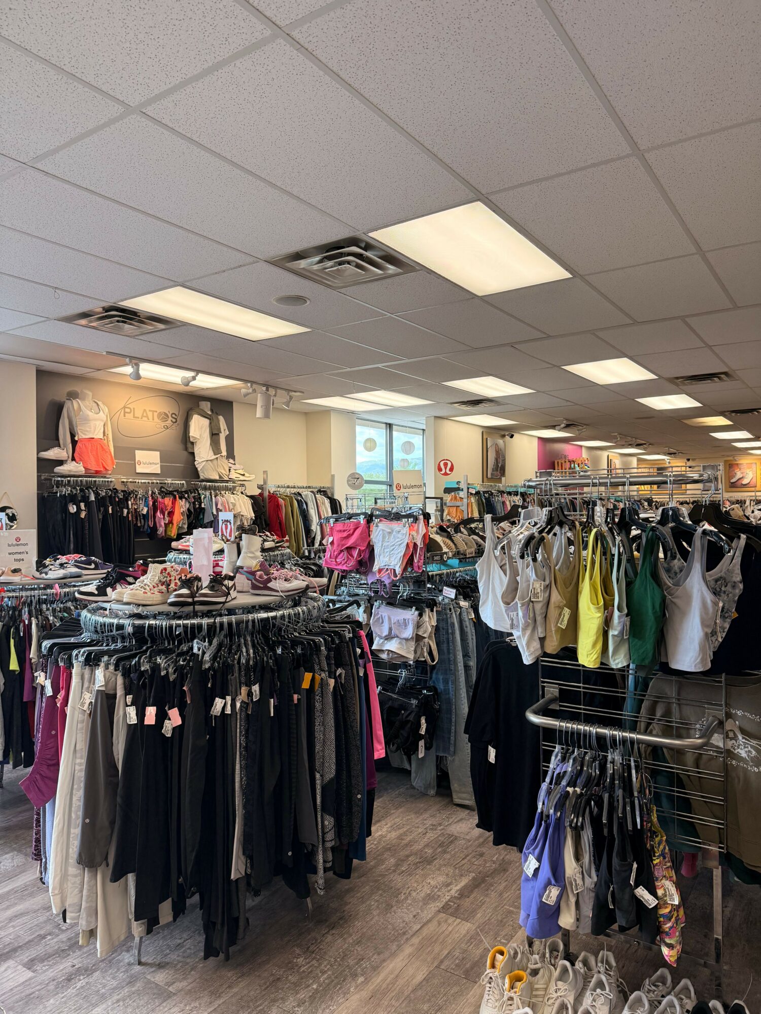 Clothing store interior with racks of clothes, tables, and shoppers browsing, viewed from the side. Bright lighting and tiled ceiling.