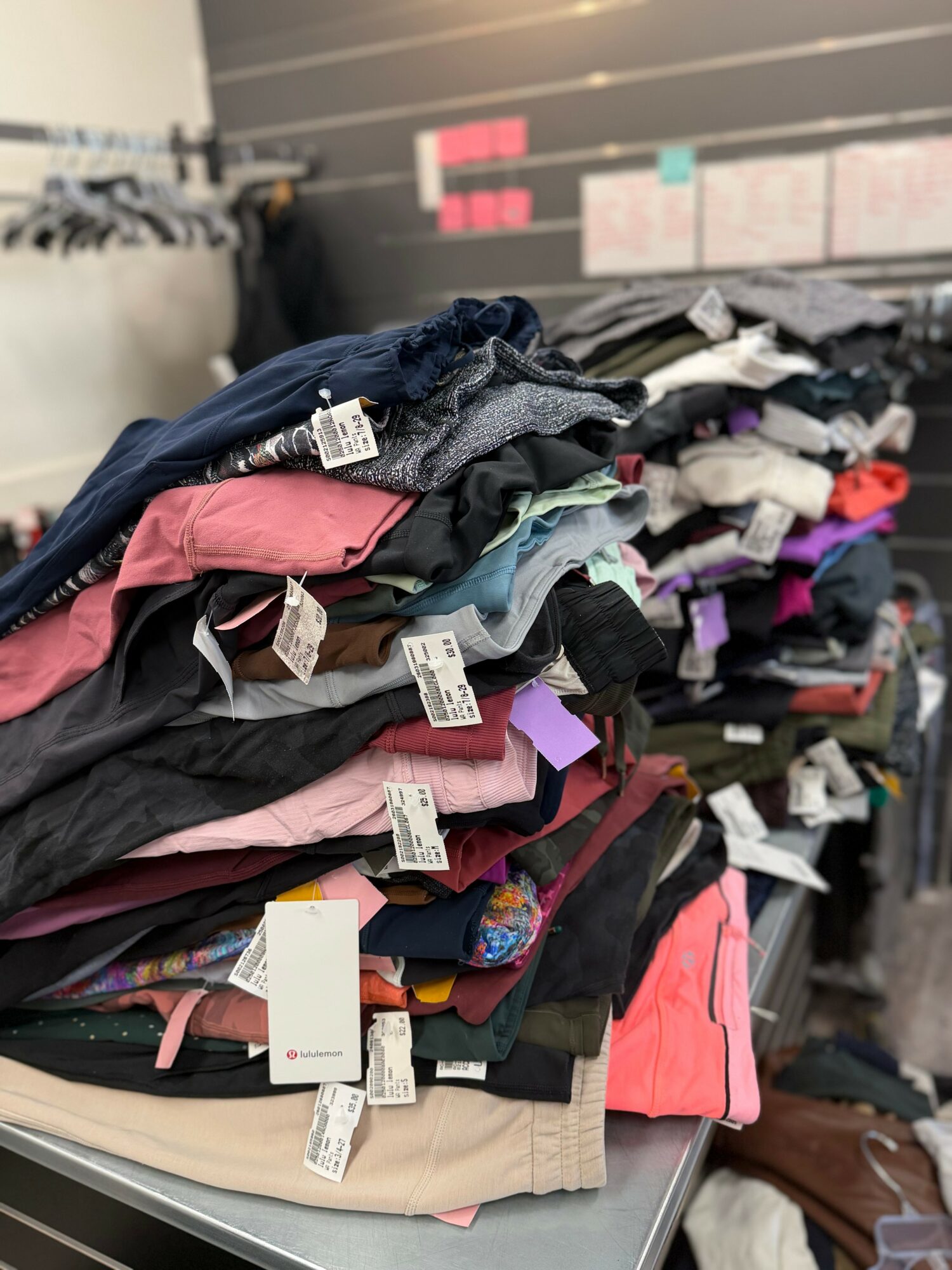 Piles of folded clothes on a table with tags, in a retail or storage setting.