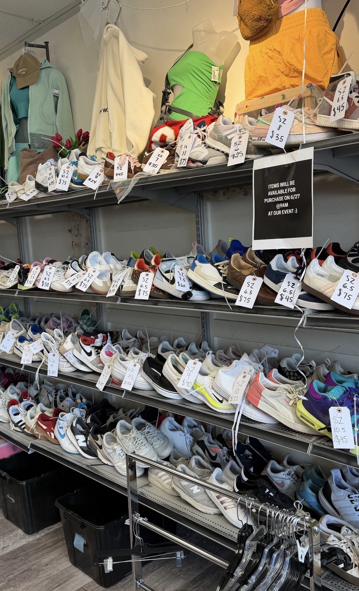 Shelves filled with various sneakers, some with tags, in a store setting, with clothing items hanging above. Black bins below.