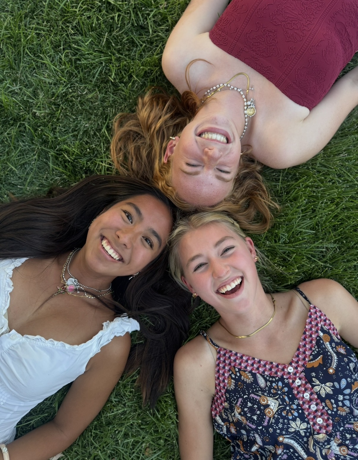 Three women lying on grass, smiling, with heads close together, wearing colorful tops and jewelry.