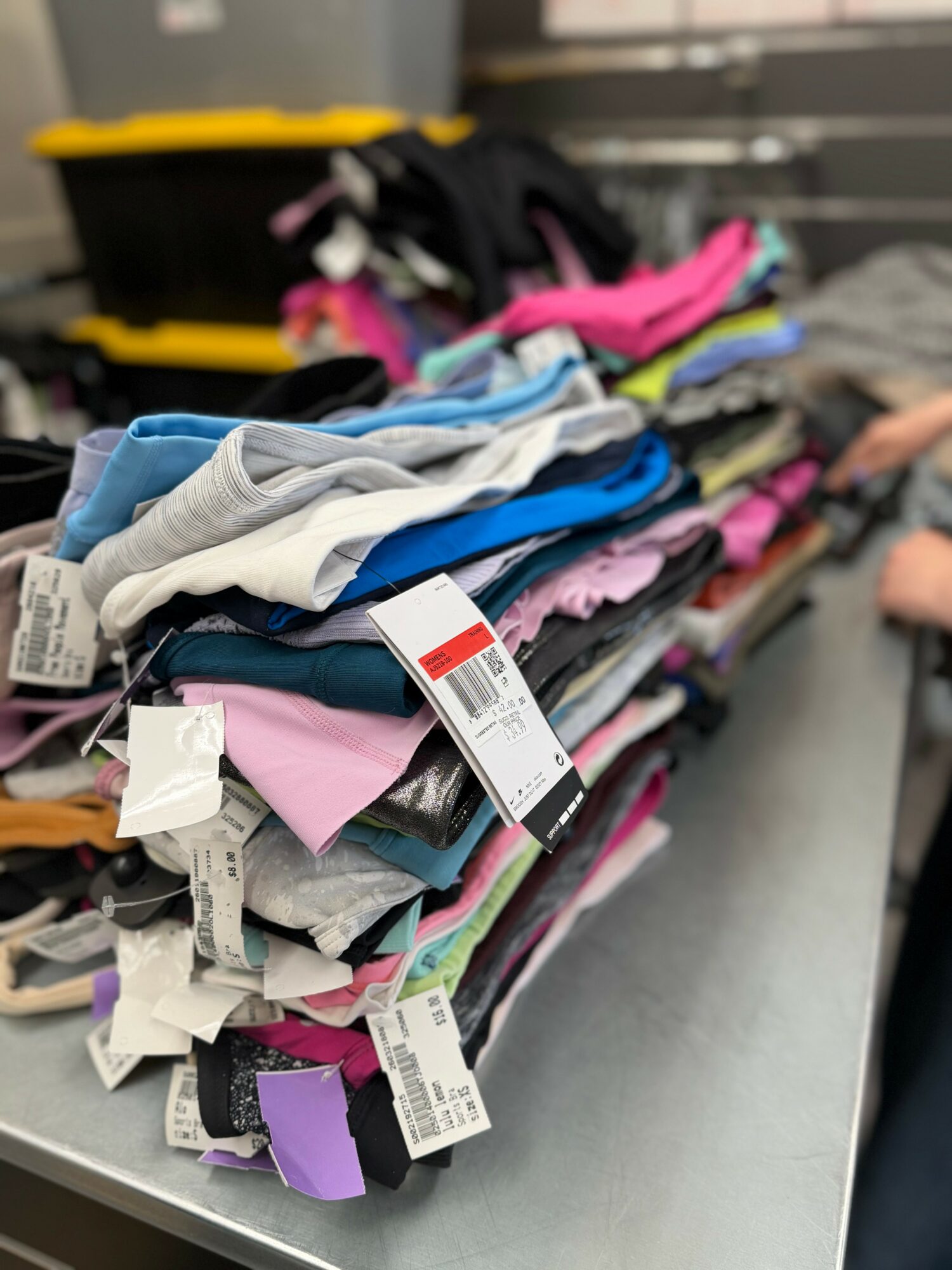 Stack of folded clothes with tags on a metal surface, with a yellow and black container in the background.