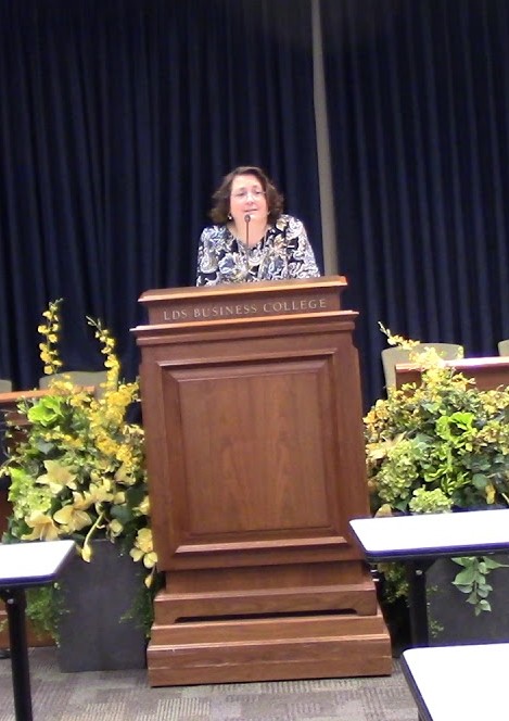 Woman standing at a wooden podium on stage with dark curtains and yellow flowers, speaking to an audience.
