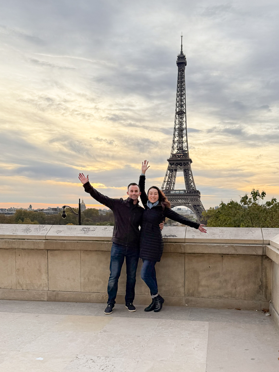 Two women stand on a balcony with the Eiffel Tower in the background, arms raised, smiling, cloudy sky above.