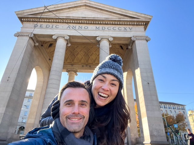 A smiling man and woman pose in front of a classical monument with columns and an inscription, under a clear blue sky.