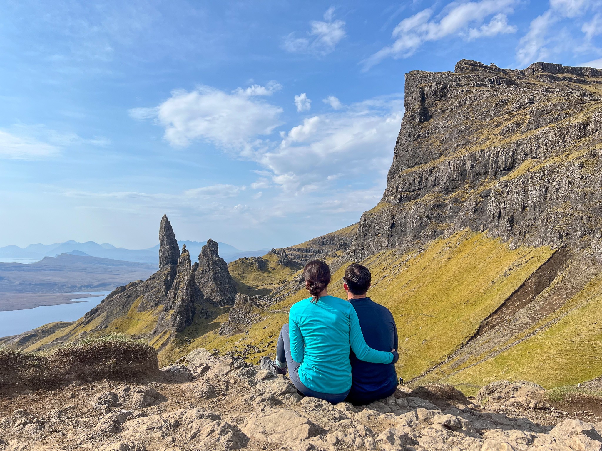 Two people sit on a rocky ledge overlooking a landscape with cliffs, mountains, and a body of water under a blue sky.
