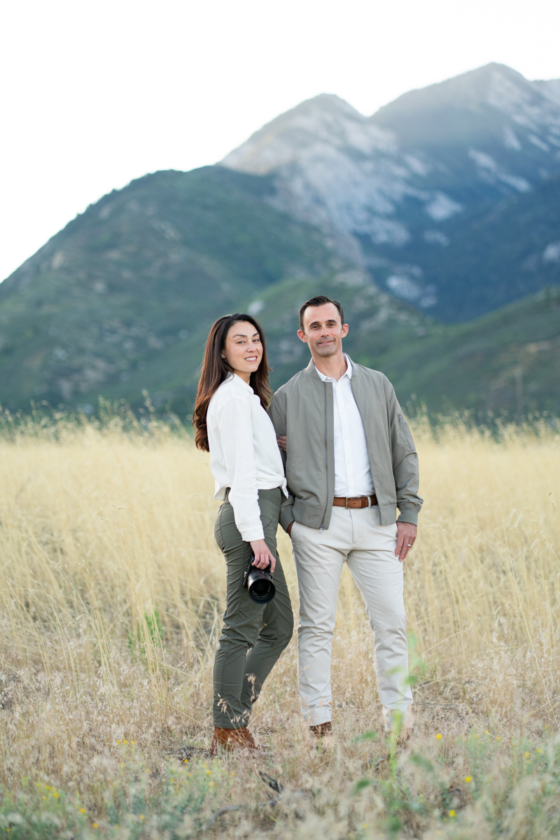 A man and woman stand in a grassy field with mountains in the background, smiling at the camera.