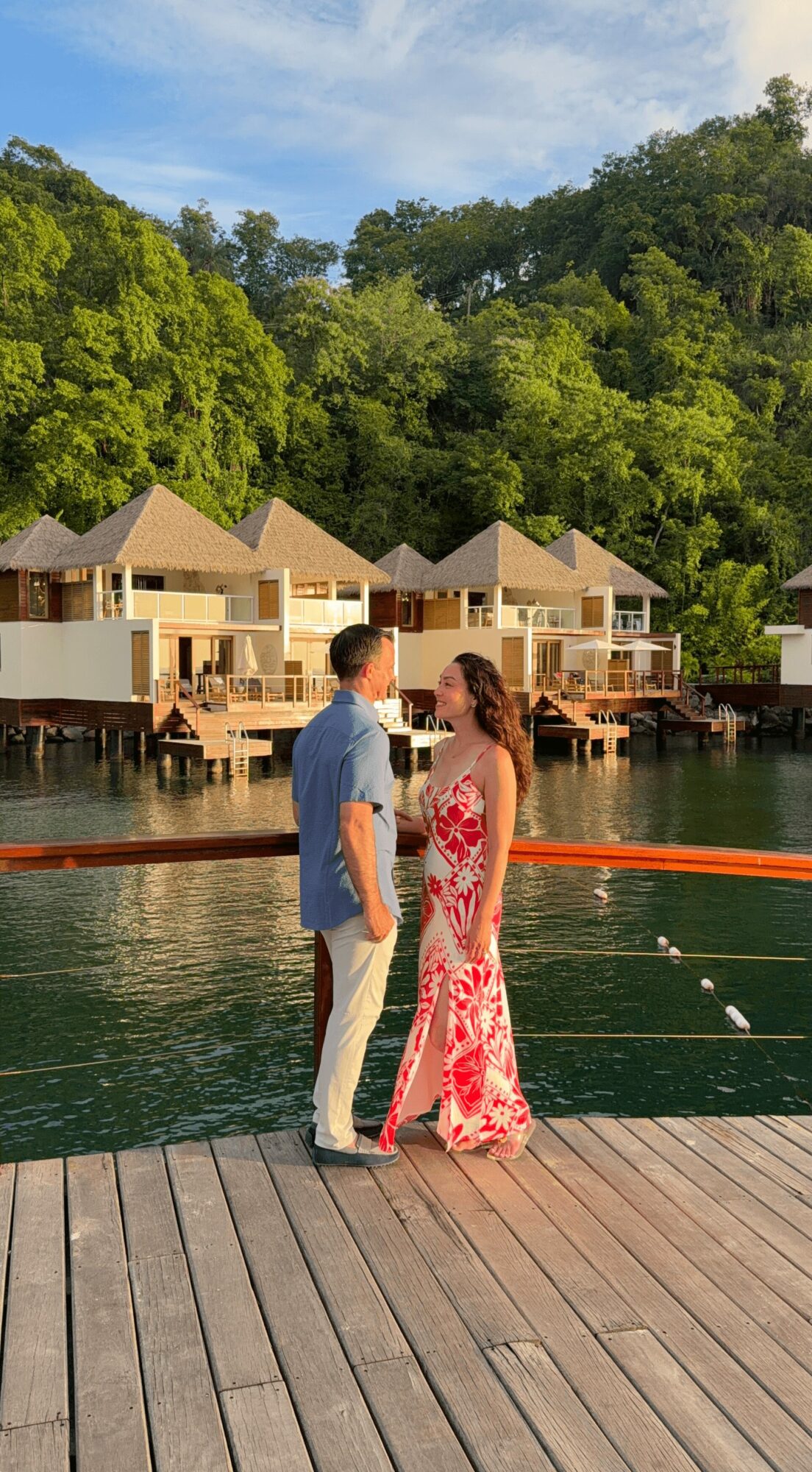A couple stands on a wooden dock near water, with overwater bungalows and green trees in the background.