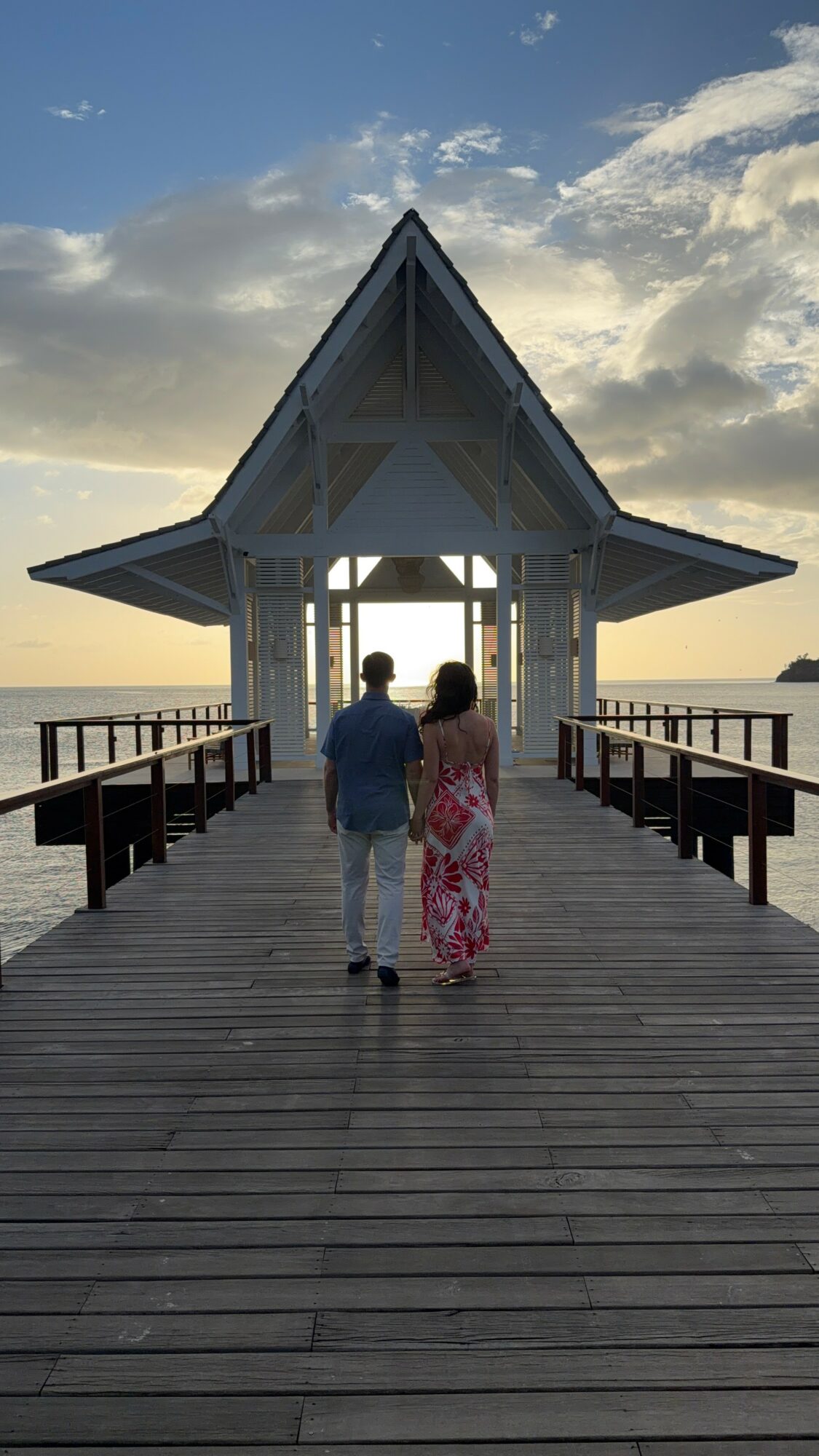 Two people walk on a wooden pier toward a pavilion over water during sunset, with clouds in the sky.