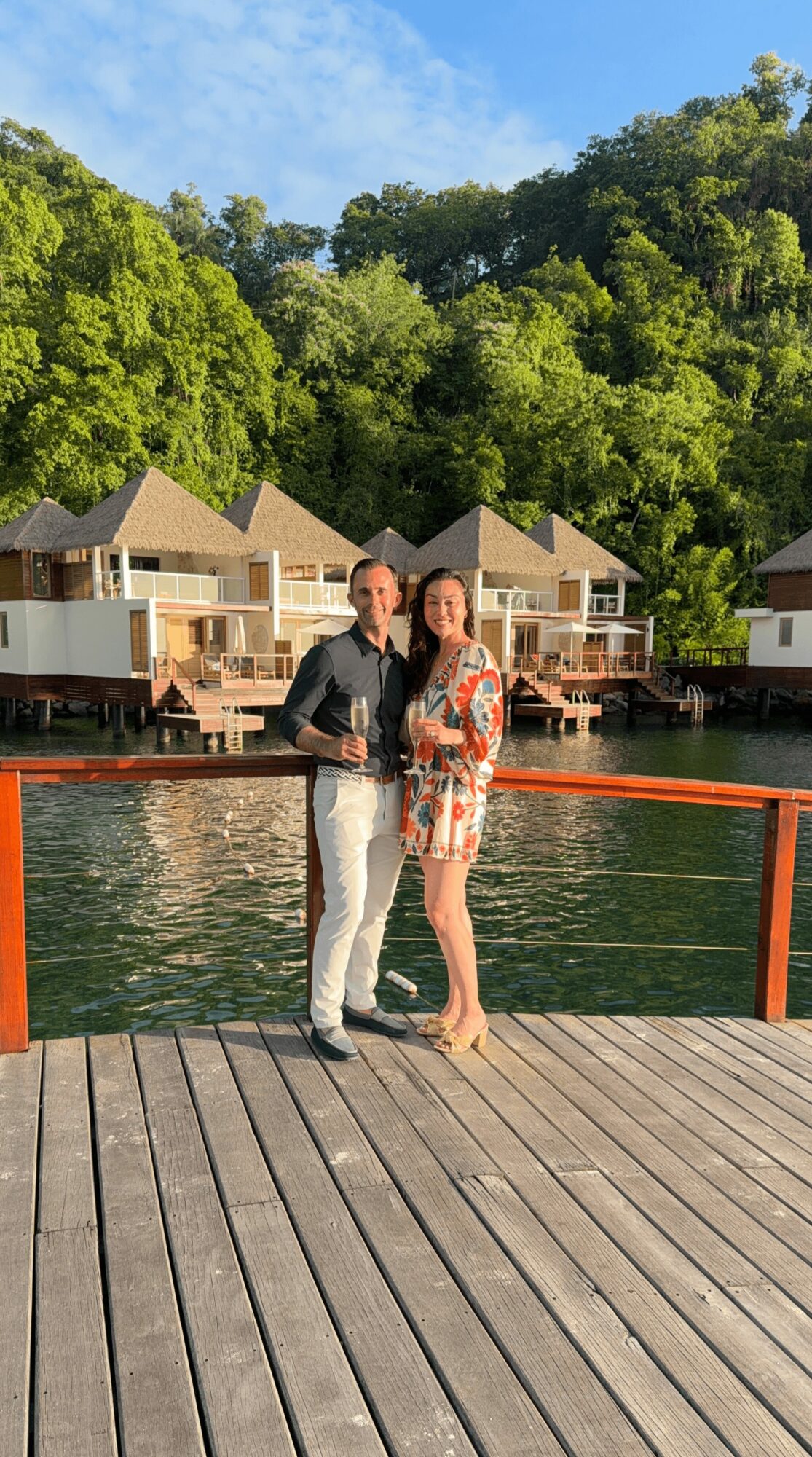 Two people standing on a dock near water, with houses and trees in the background, smiling and holding drinks.