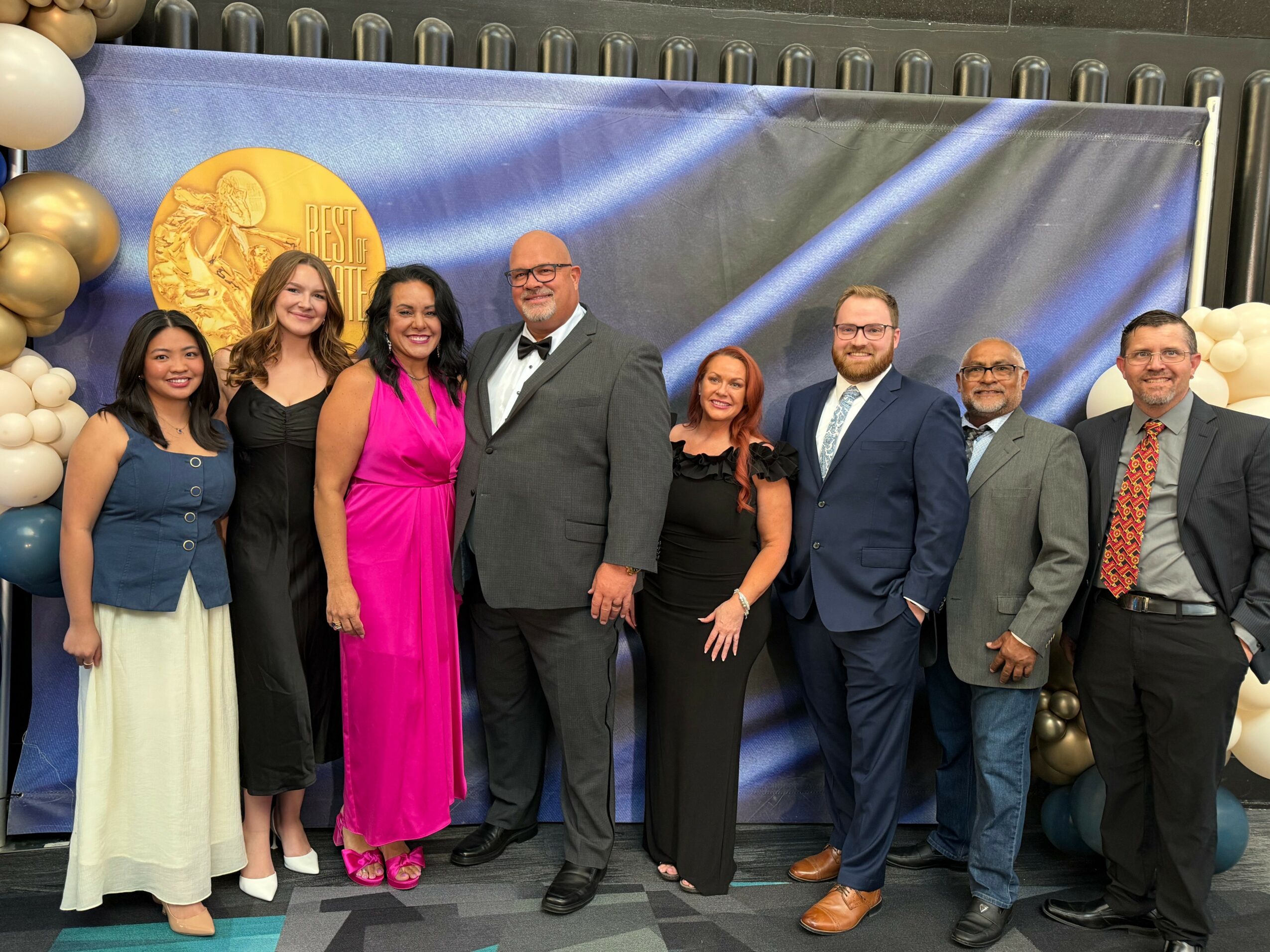Group of nine people standing in front of a blue backdrop with gold balloons on the left side, smiling for a photo.