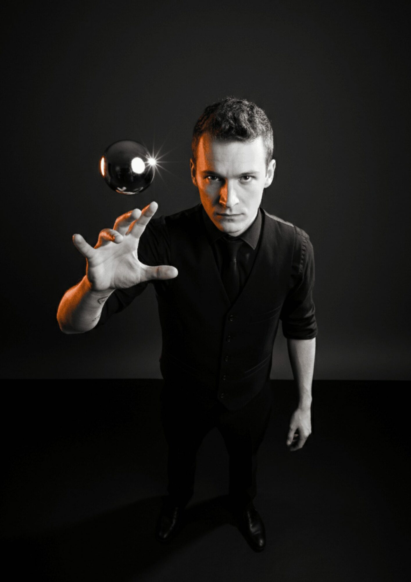 Young man with short hair and dark clothing reaches out towards a floating shiny sphere against a dark background.