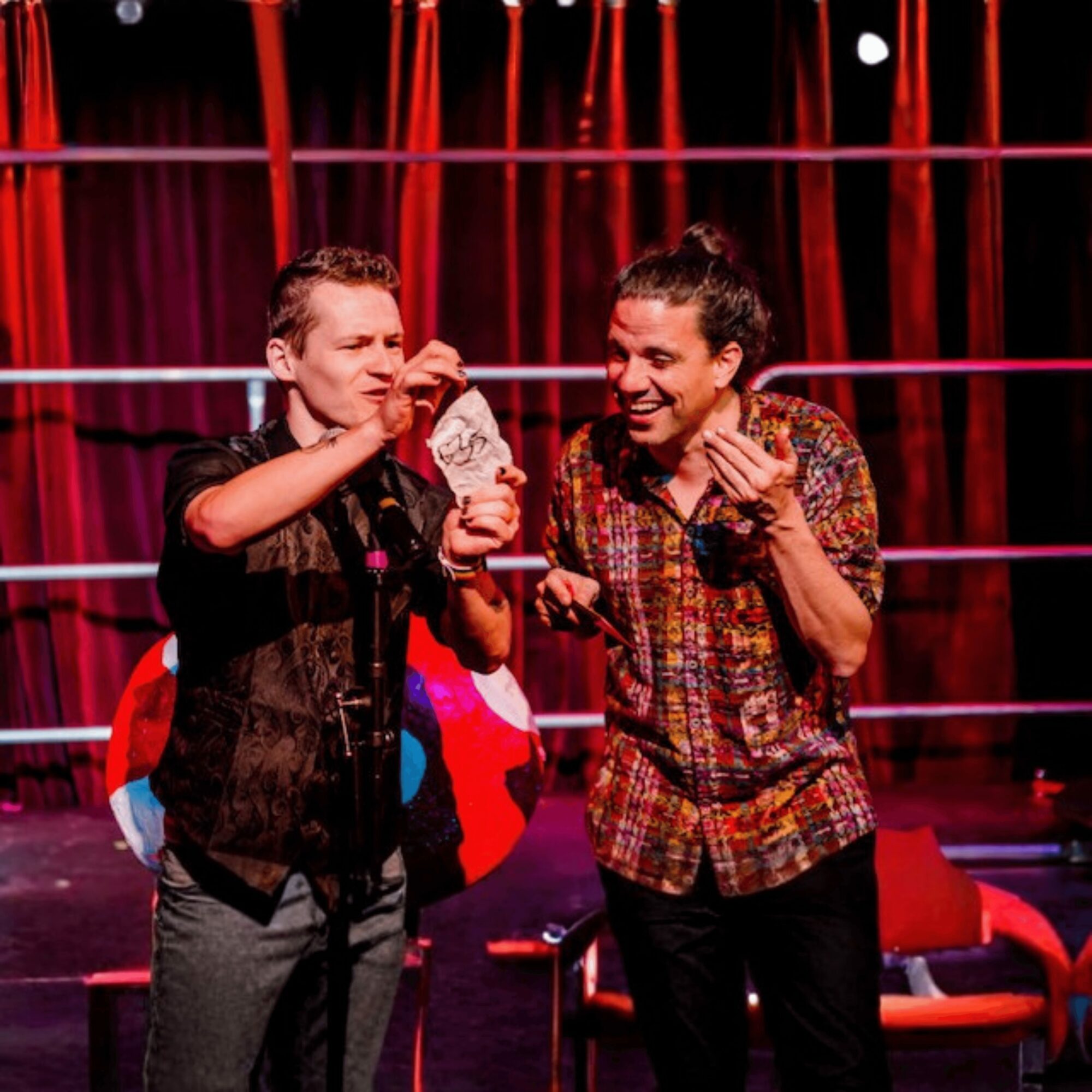 Two people laughing and talking on stage with red curtains and boxing ring ropes in background.