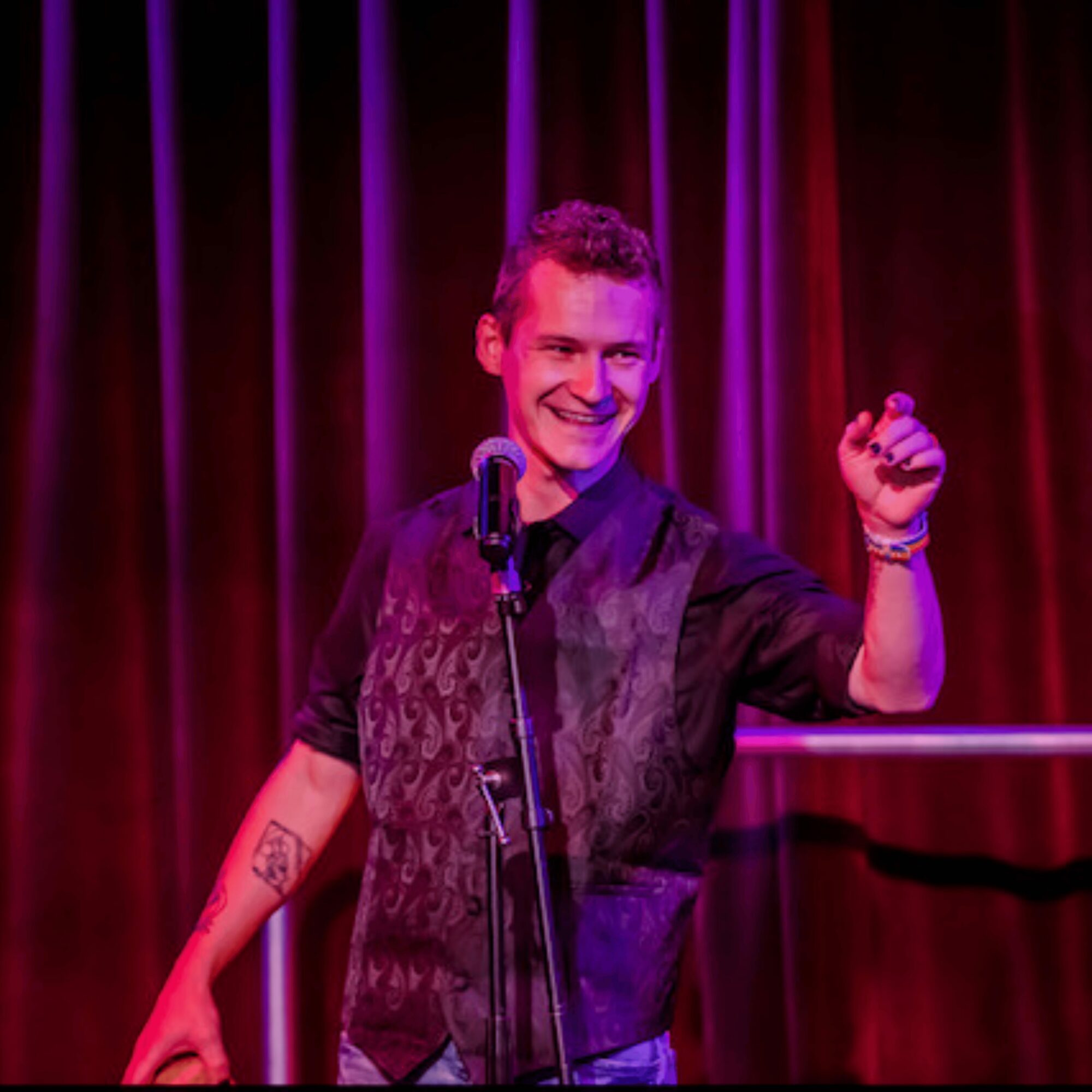 Person on stage with a microphone, smiling, raising a fist, red curtain background.