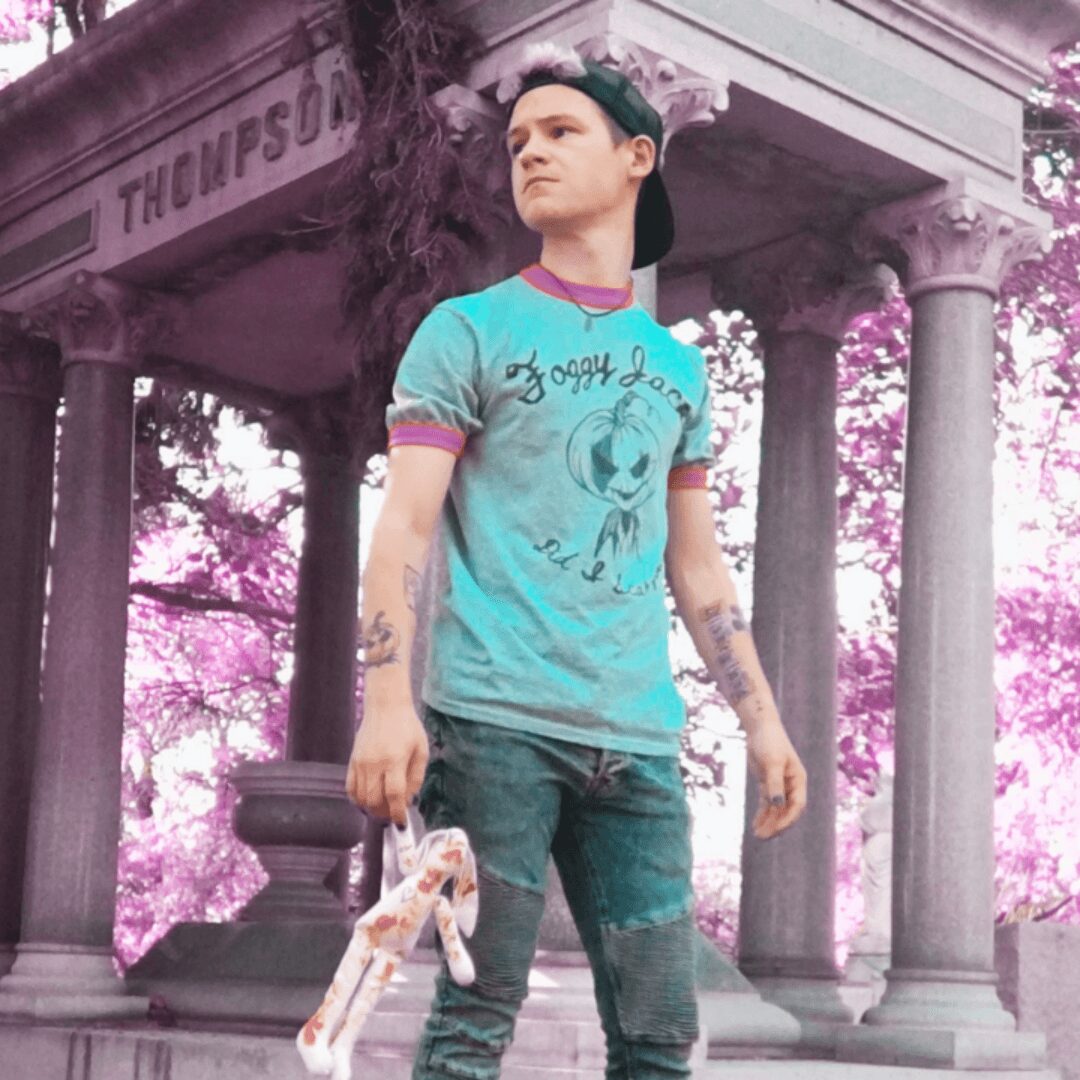 Young man standing in front of a classical structure with columns, holding a skateboard, with pink trees in the background.
