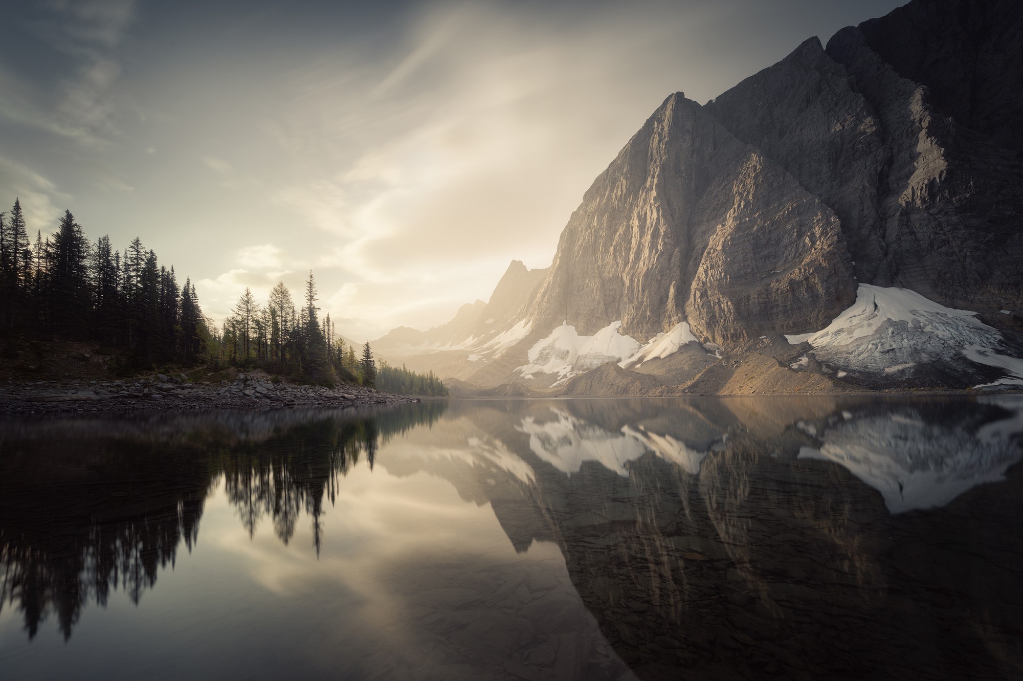 Mountain landscape with a calm lake reflecting mountains and trees, cloudy sky, and snow patches on the mountainside.