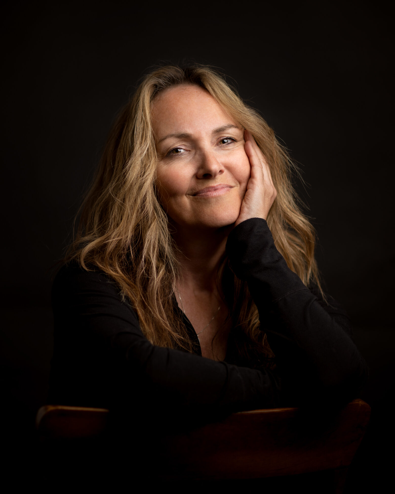 Woman with wavy blonde hair resting her face on her hand, smiling softly, wearing a black top, dark background.