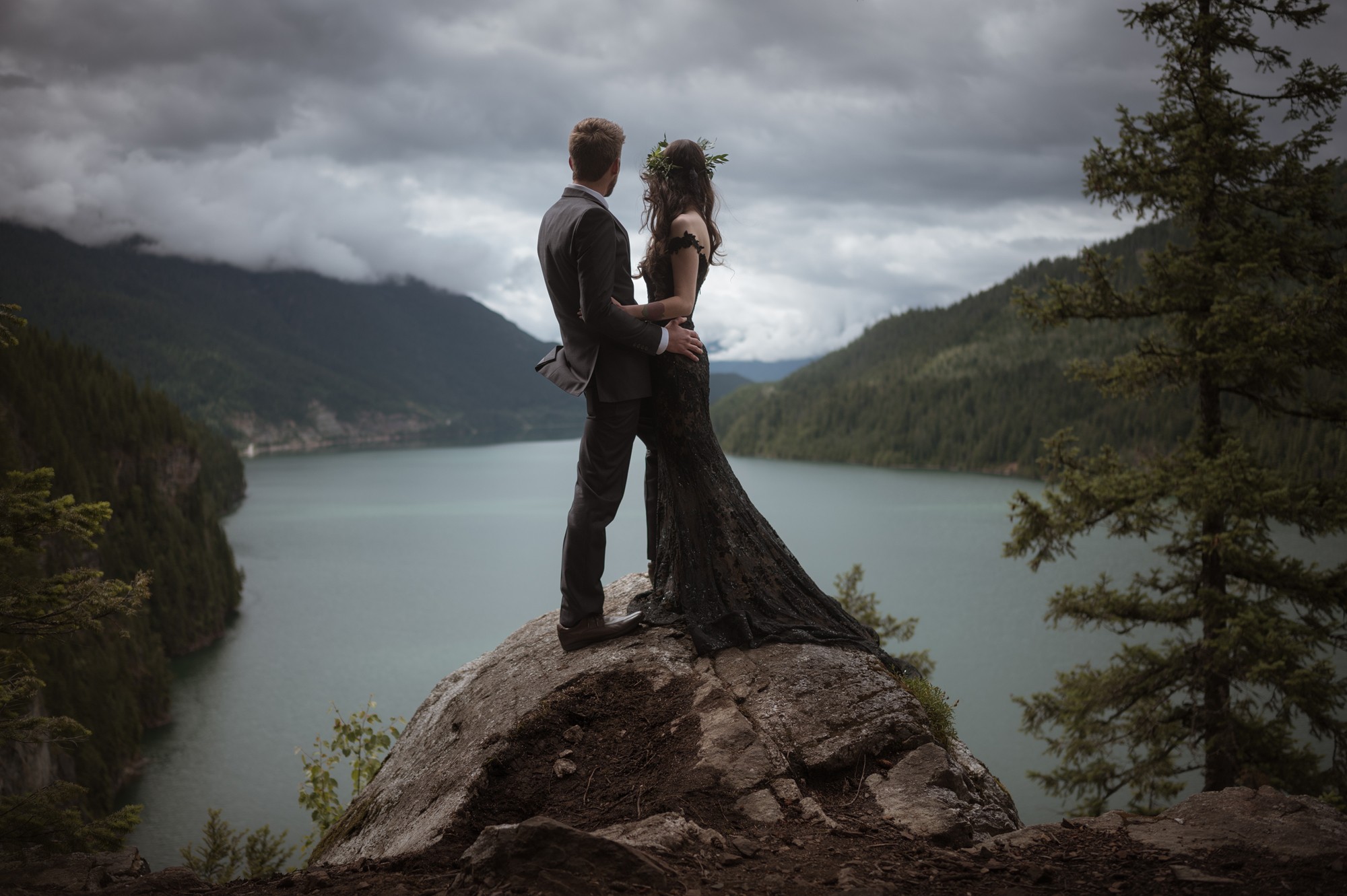 A couple stands on a large rock overlooking a lake with mountains and cloudy sky in the background.