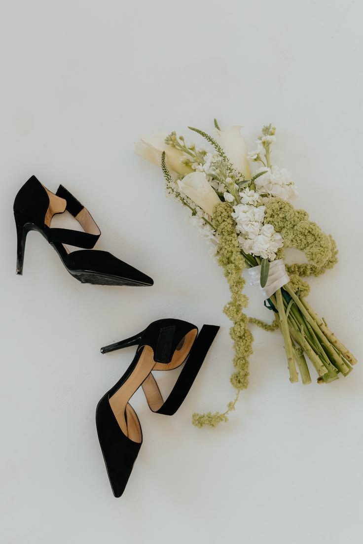 Pair of black high-heeled shoes and a bouquet of white and green flowers on a white background.