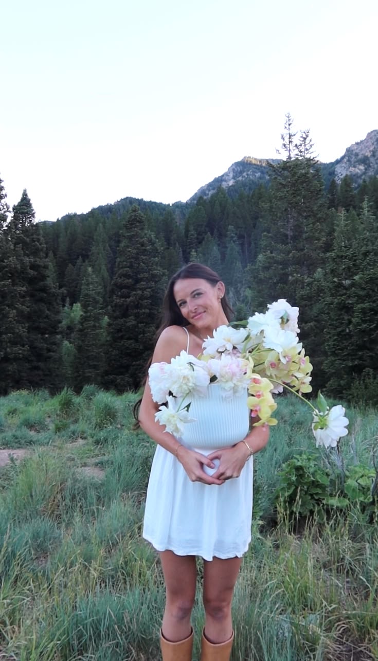 Woman in white dress holding a large bouquet of white flowers outdoors with mountains and trees in background