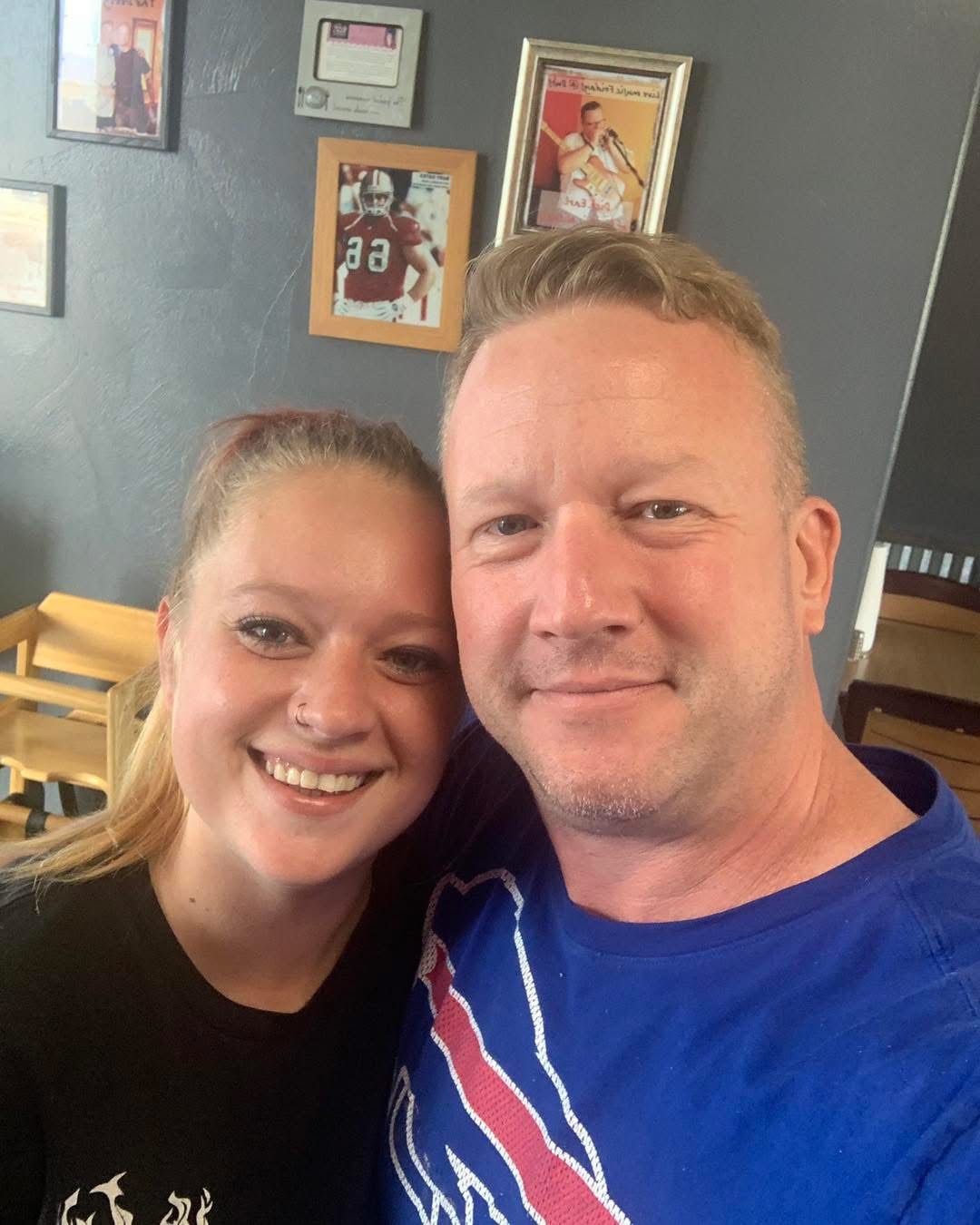Two people smiling for a selfie indoors, with framed pictures on the wall behind them.