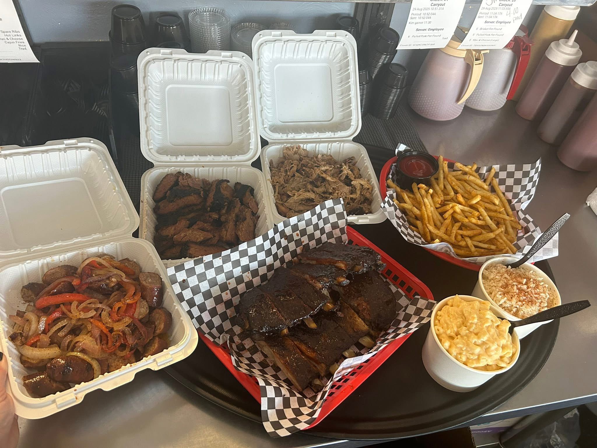 Takeout food containers with ribs, pasta, and fries on a table, with drinks in the background.