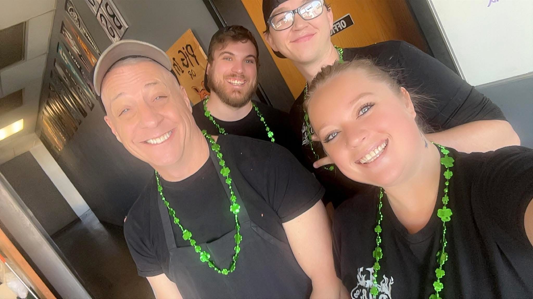 Four smiling people wearing green bead necklaces taking a selfie indoors.