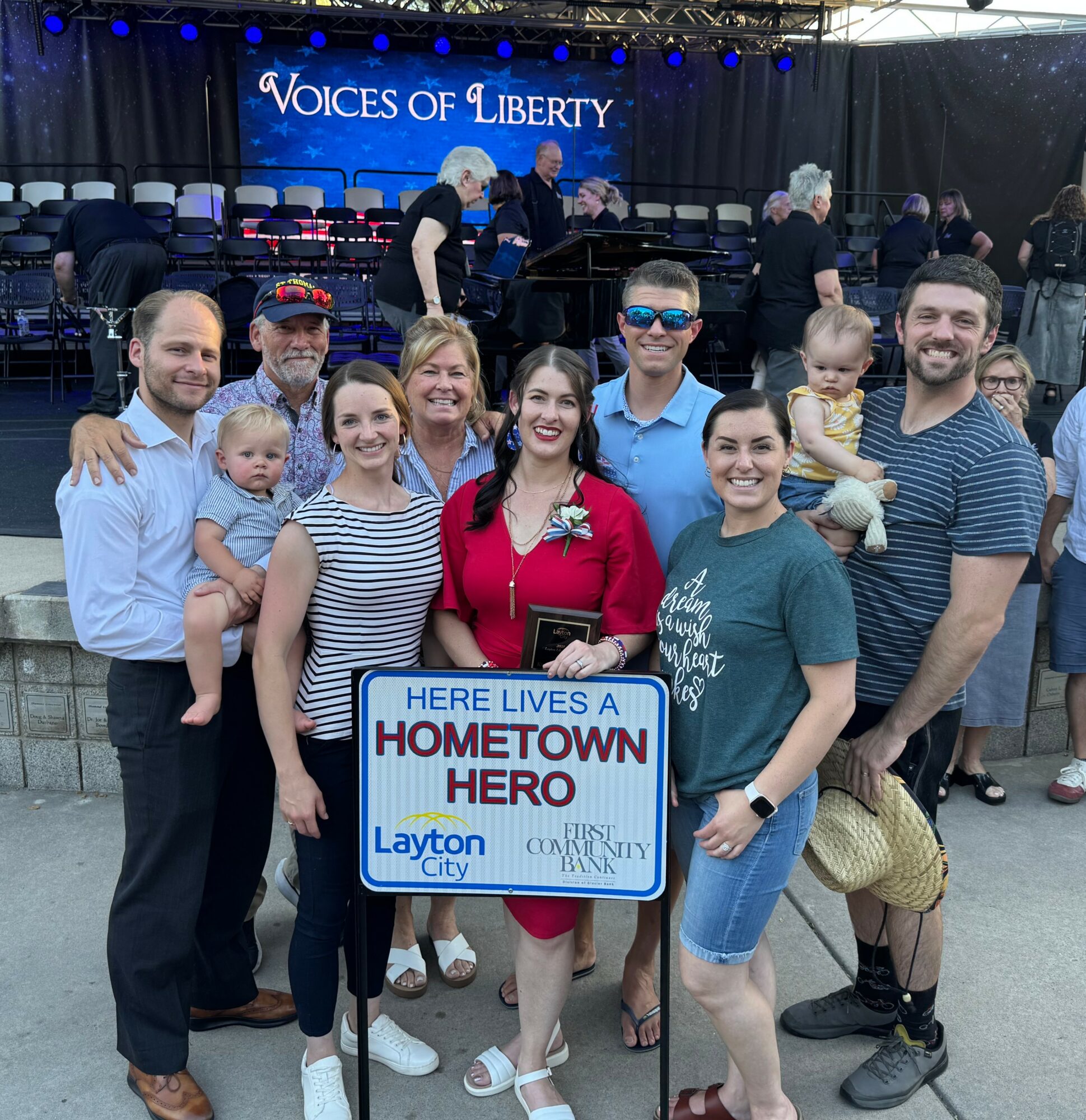 Group of people, including children, holding a sign that reads 'Here lives a hometown hero,' at an outdoor event with a stage in the background.