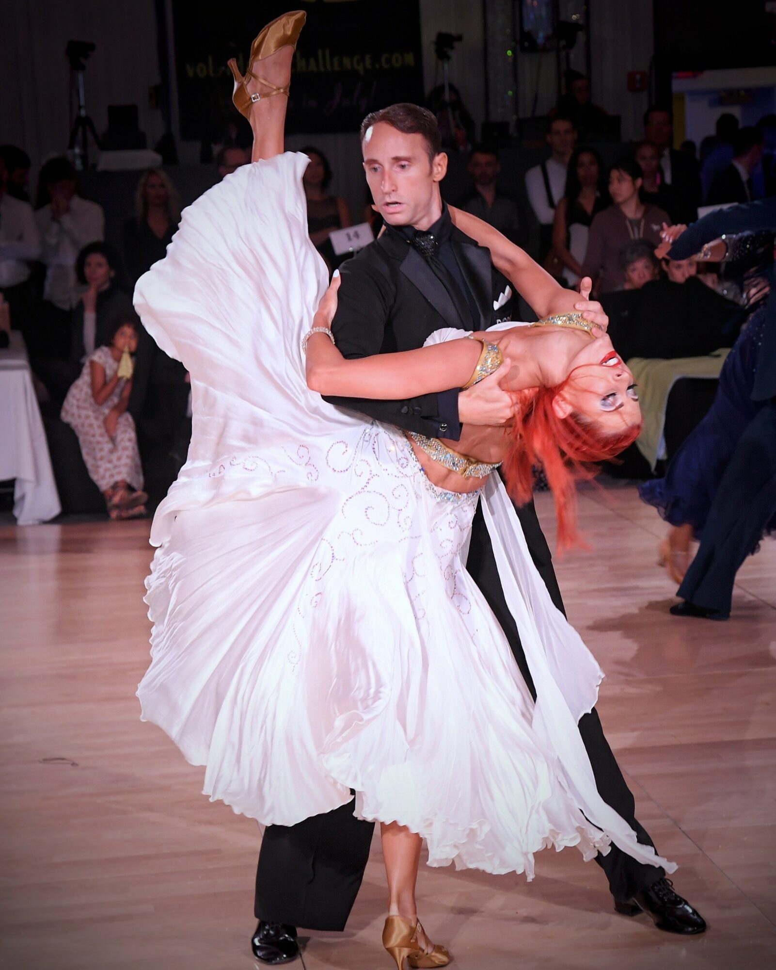 A couple dancing ballroom, the man in a black suit and the woman in a white dress, with an audience in the background.