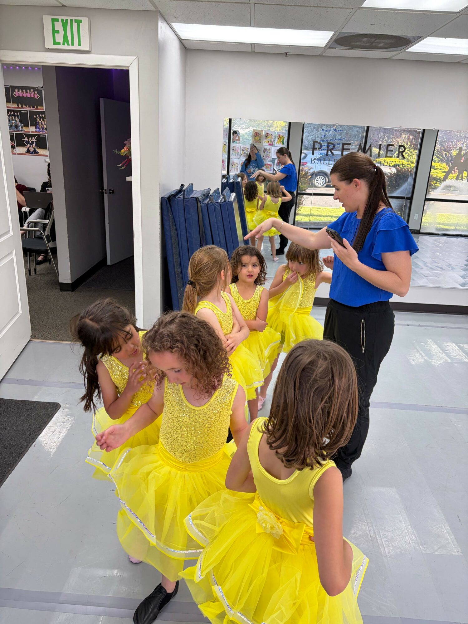 Children in yellow dresses and adults in blue and black shirts in a hallway, some sitting and others standing, with a blue curtain and posters in the background.