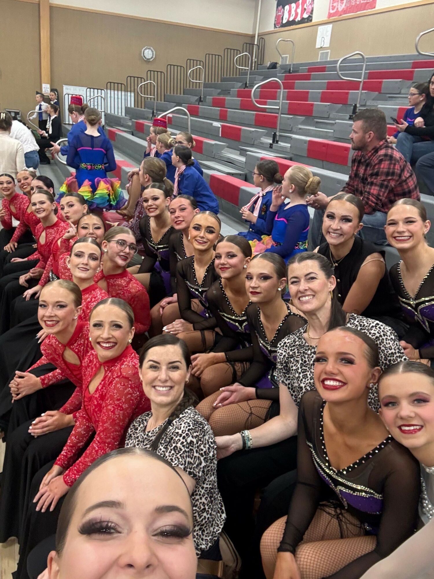 Group of young women in colorful costumes sitting on bleachers, smiling and posing for a photo indoors.