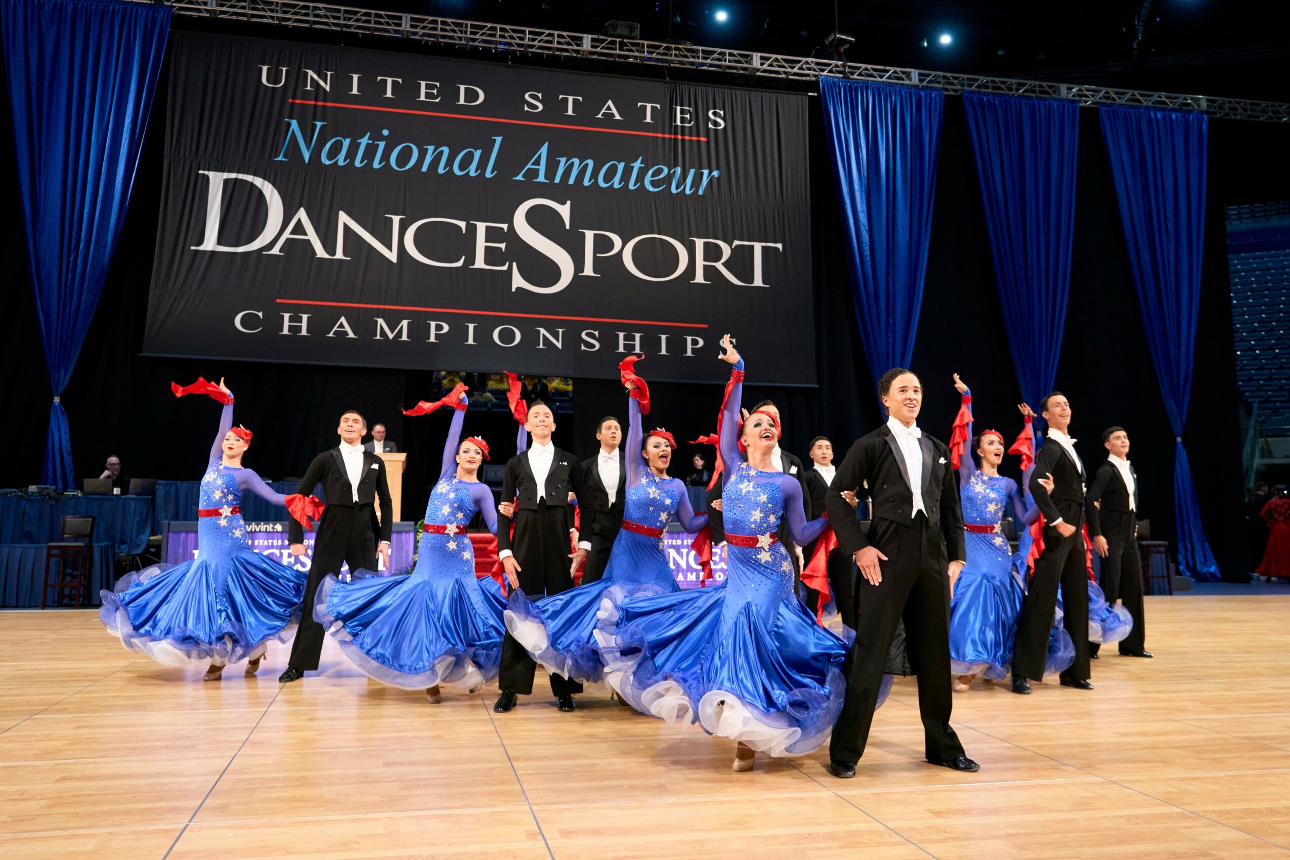 Group of dancers in blue dresses and black suits performing on stage at dance championships, with a large banner behind them.
