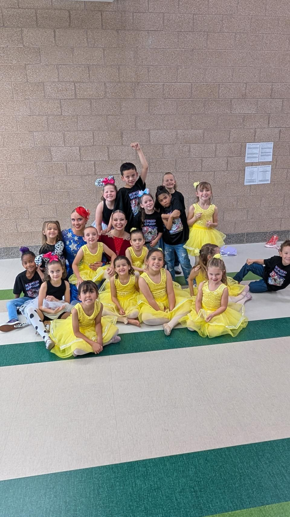 Group of children in yellow costumes and others in black costumes, sitting and standing on a green and white striped floor, against a brick wall.