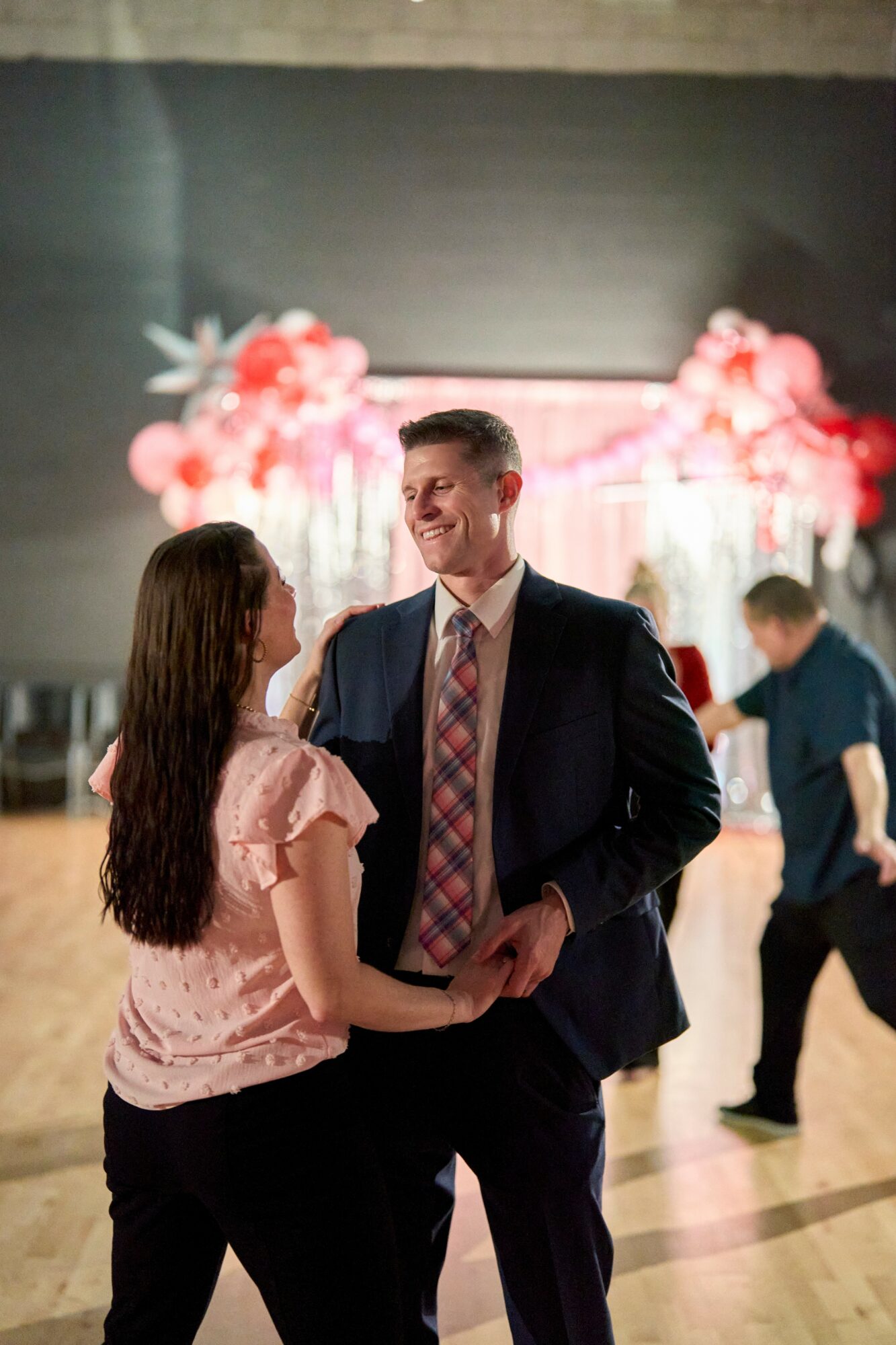A man and woman dance together at an event with pink balloon decorations in the background.
