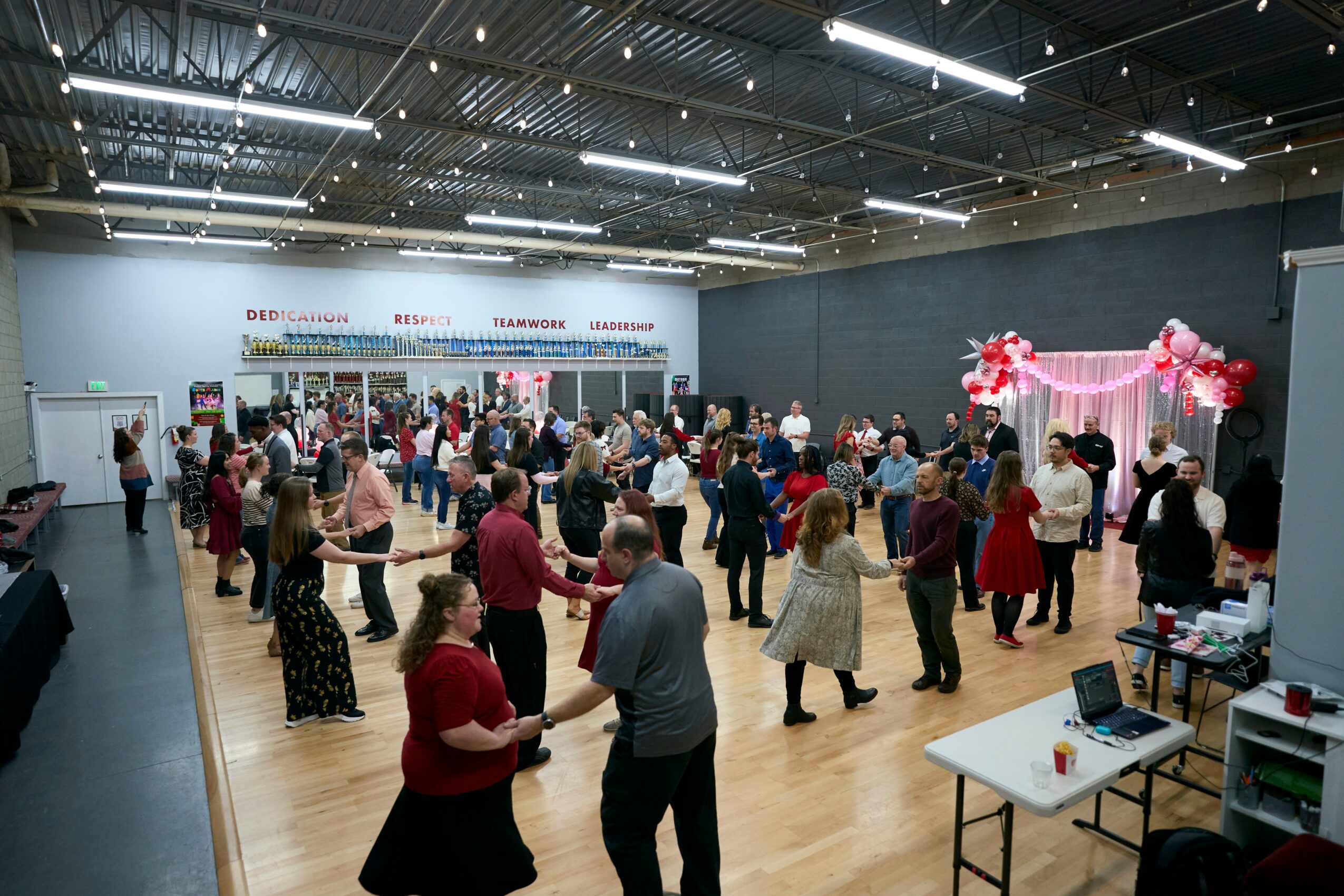 People gather in a large indoor space decorated with pink flowers and ribbons for an event or celebration.
