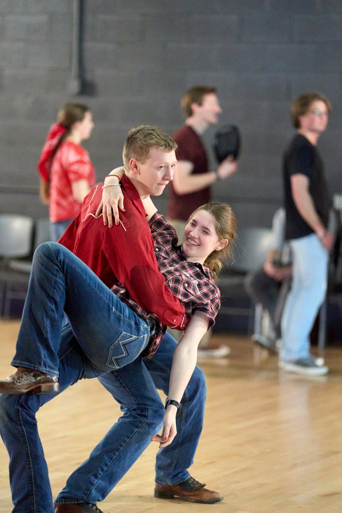 Two people dancing in a gymnasium, one lifting the other, with others in the background. Bright lighting and wooden floor.