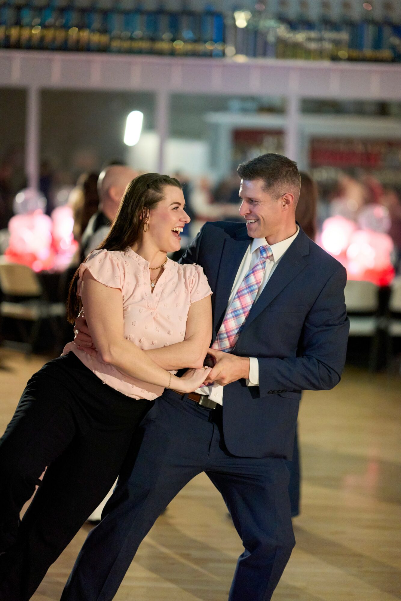 A man and woman dance together, smiling and holding hands, in a lively indoor setting with blurred background.