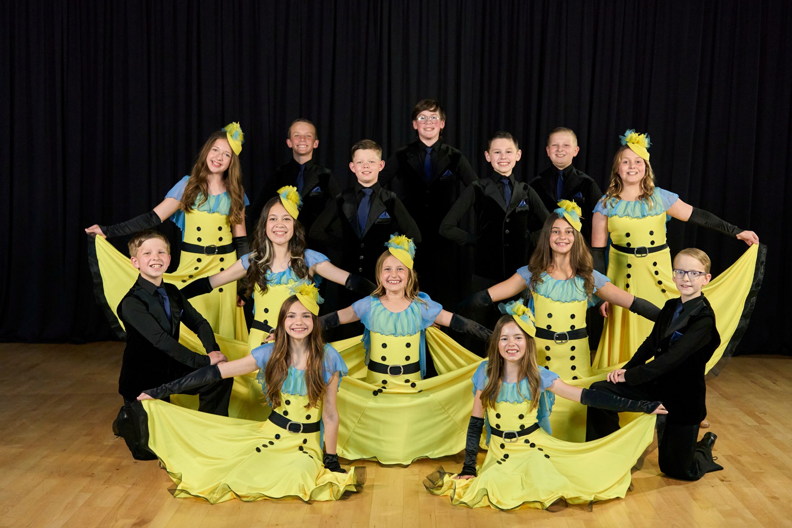 Group of children in colorful costumes posing on stage with black curtain background.