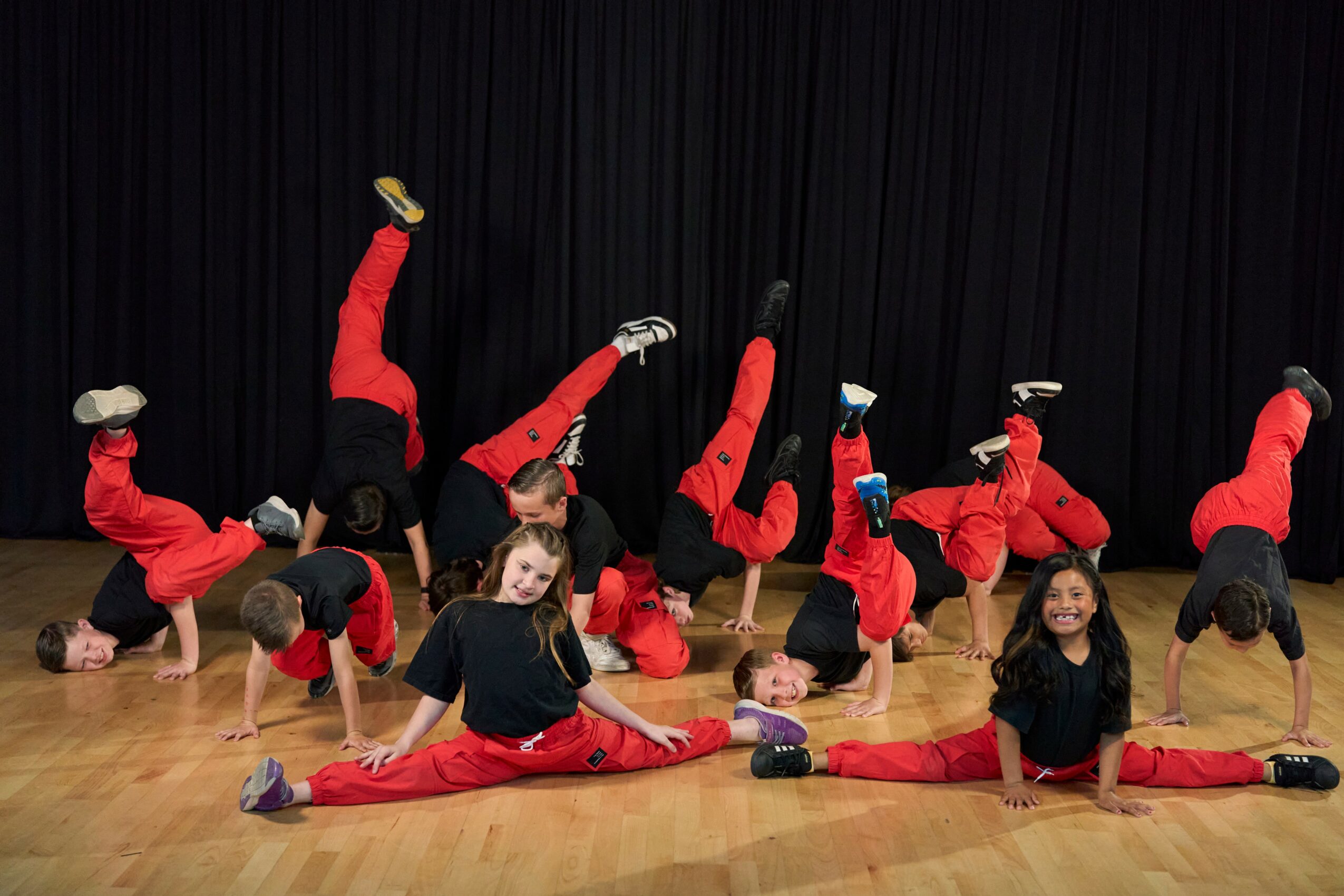 Group of children in red and black outfits performing dance moves on stage with black curtains behind them.