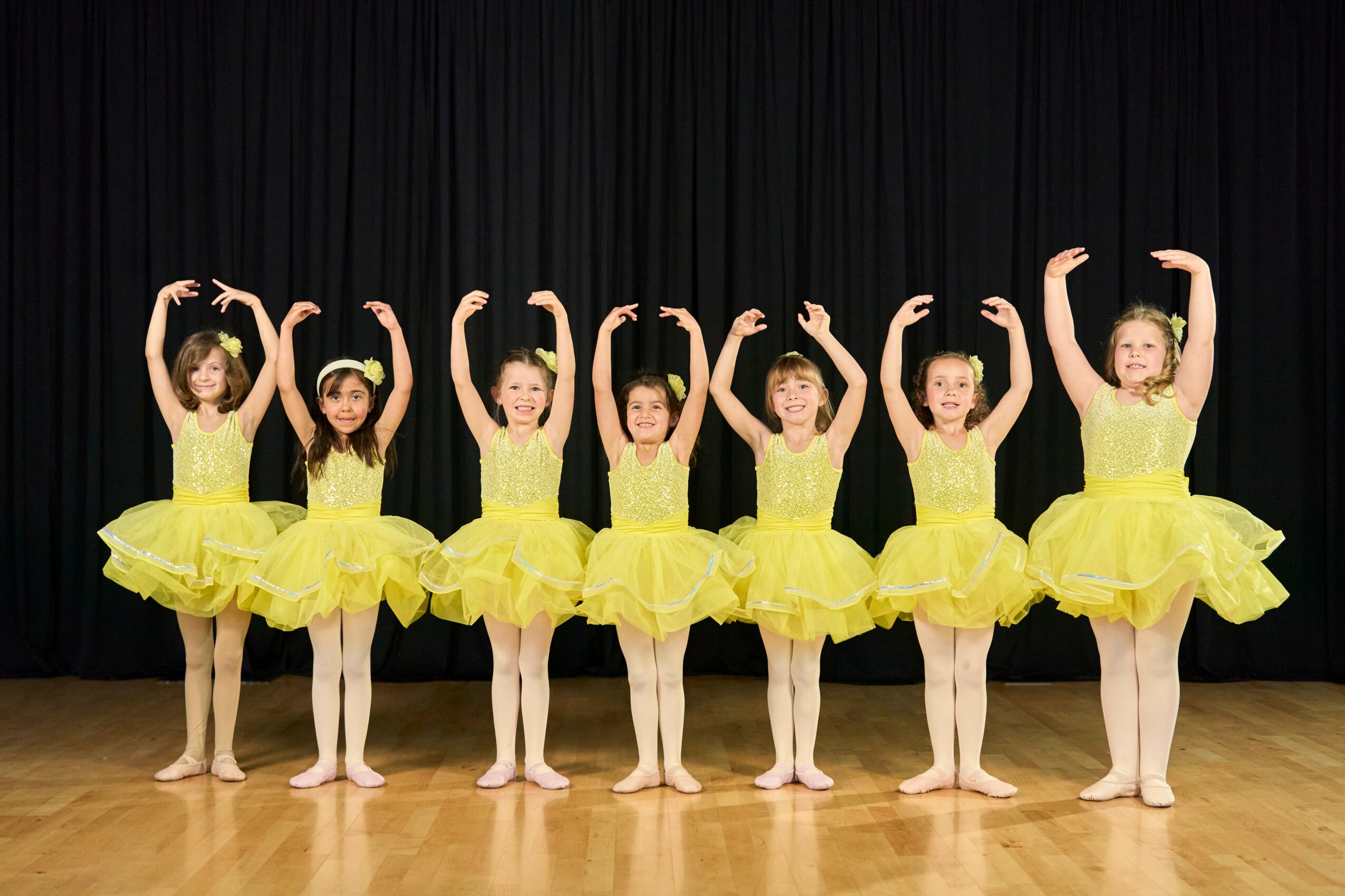 Seven young ballet dancers in yellow tutus and tights pose with arms raised in front of a black curtain.