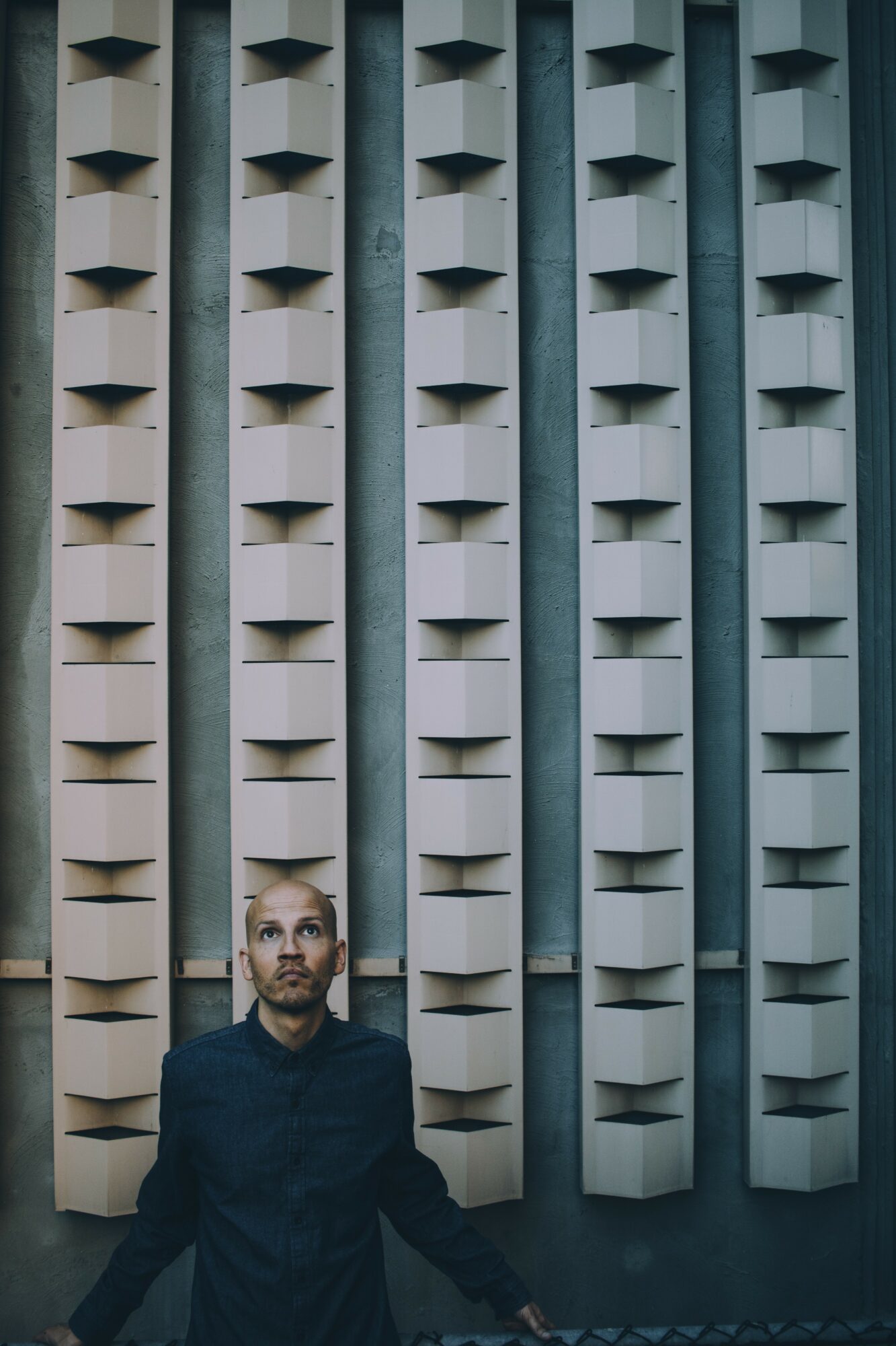 Man with a beard and dark shirt stands in front of a modern building facade with vertical, rectangular windows.